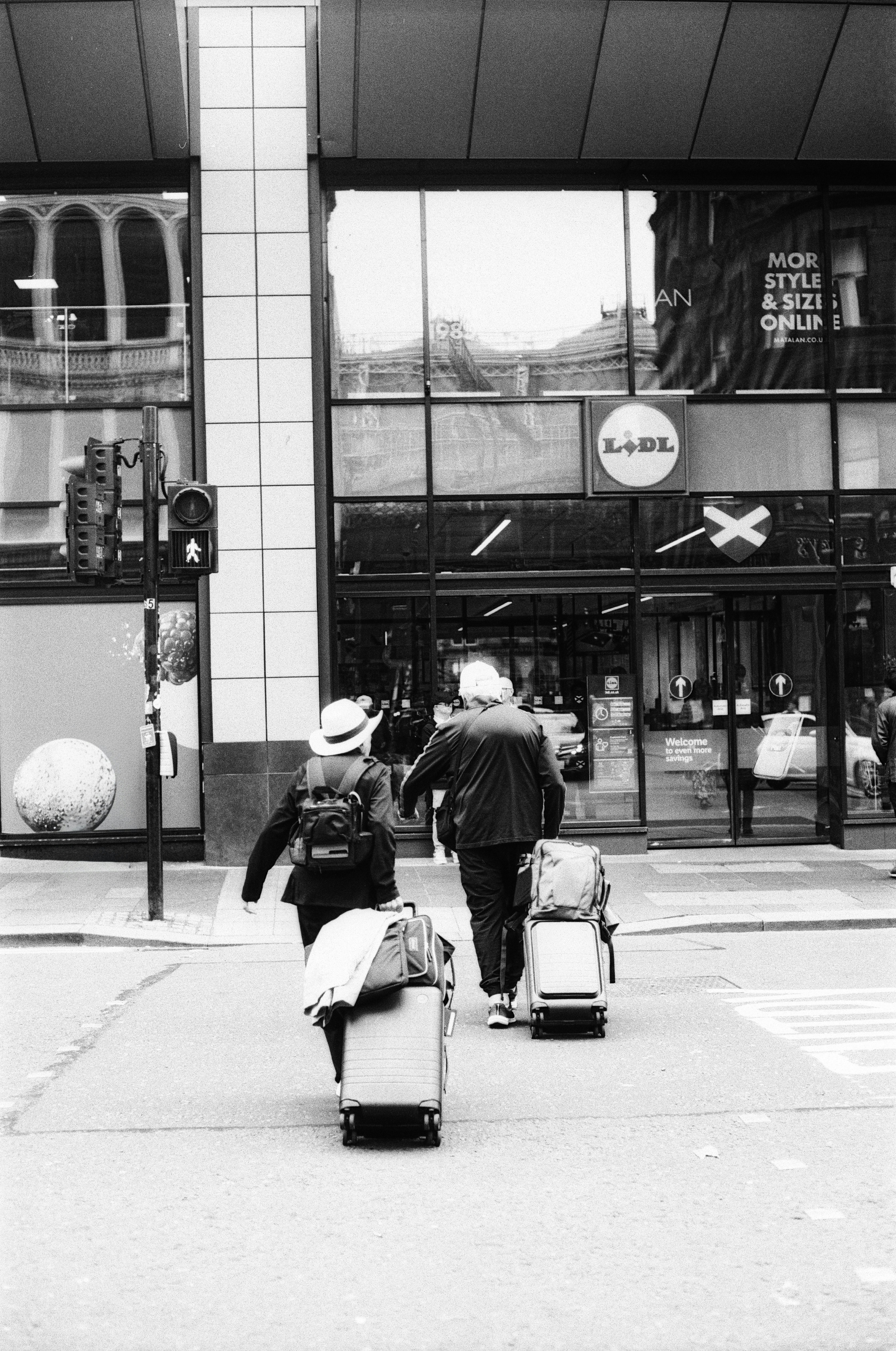 Two people with rolling suitcases crossing a city street in front of a store