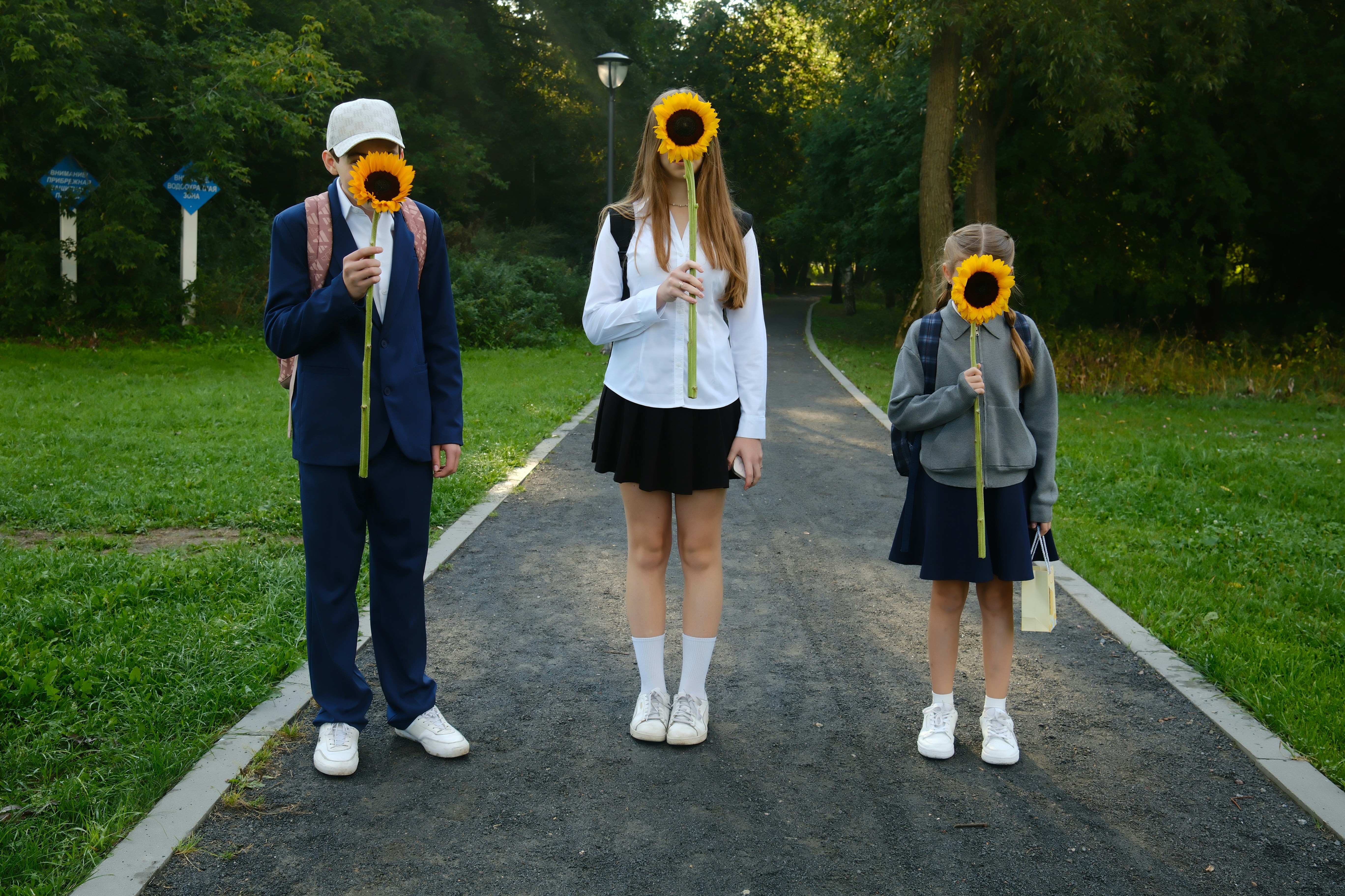 Three students holding sunflowers in front of their faces on a park path