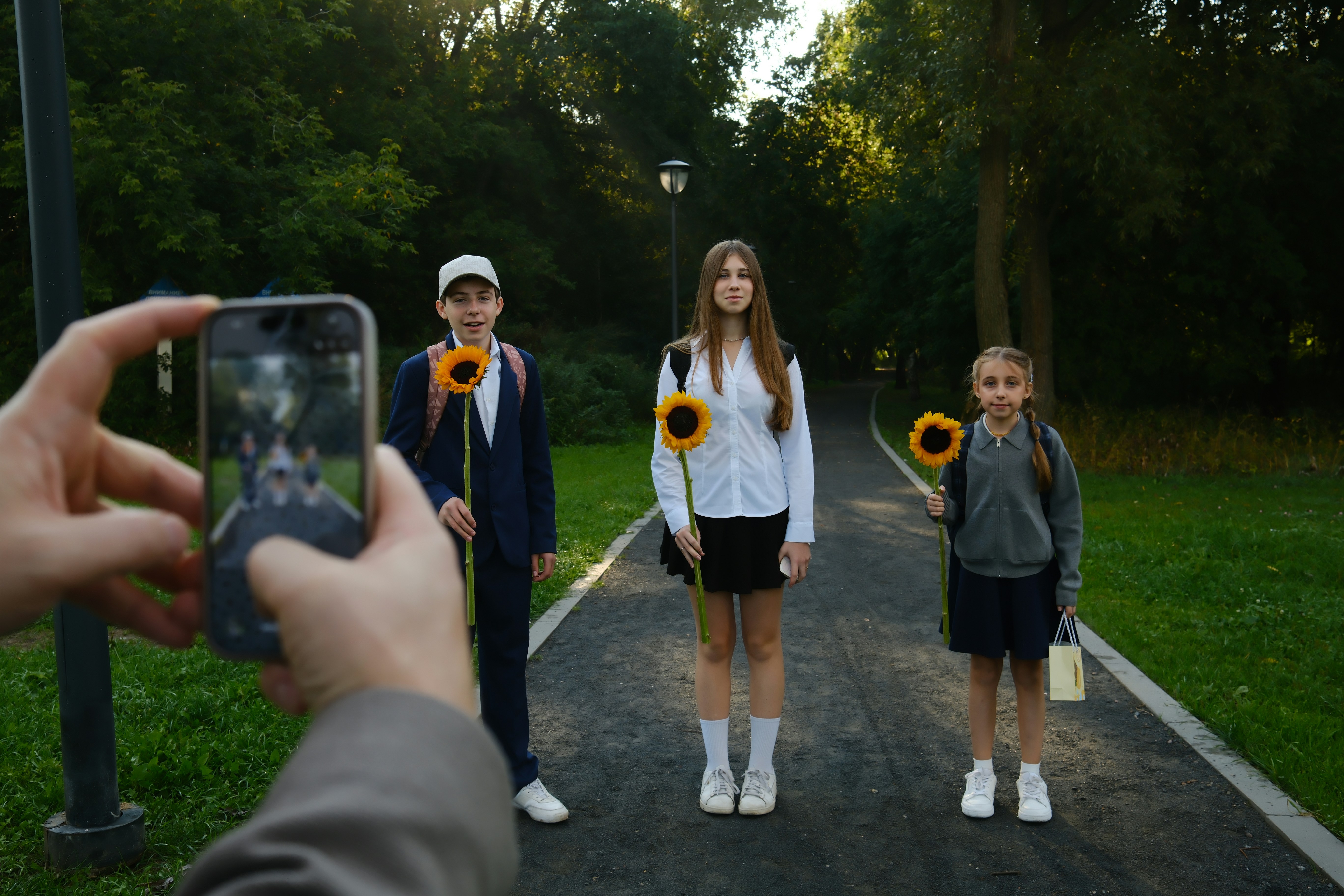 A person holding a phone photographing three children standing on a park path holding sunflowers
