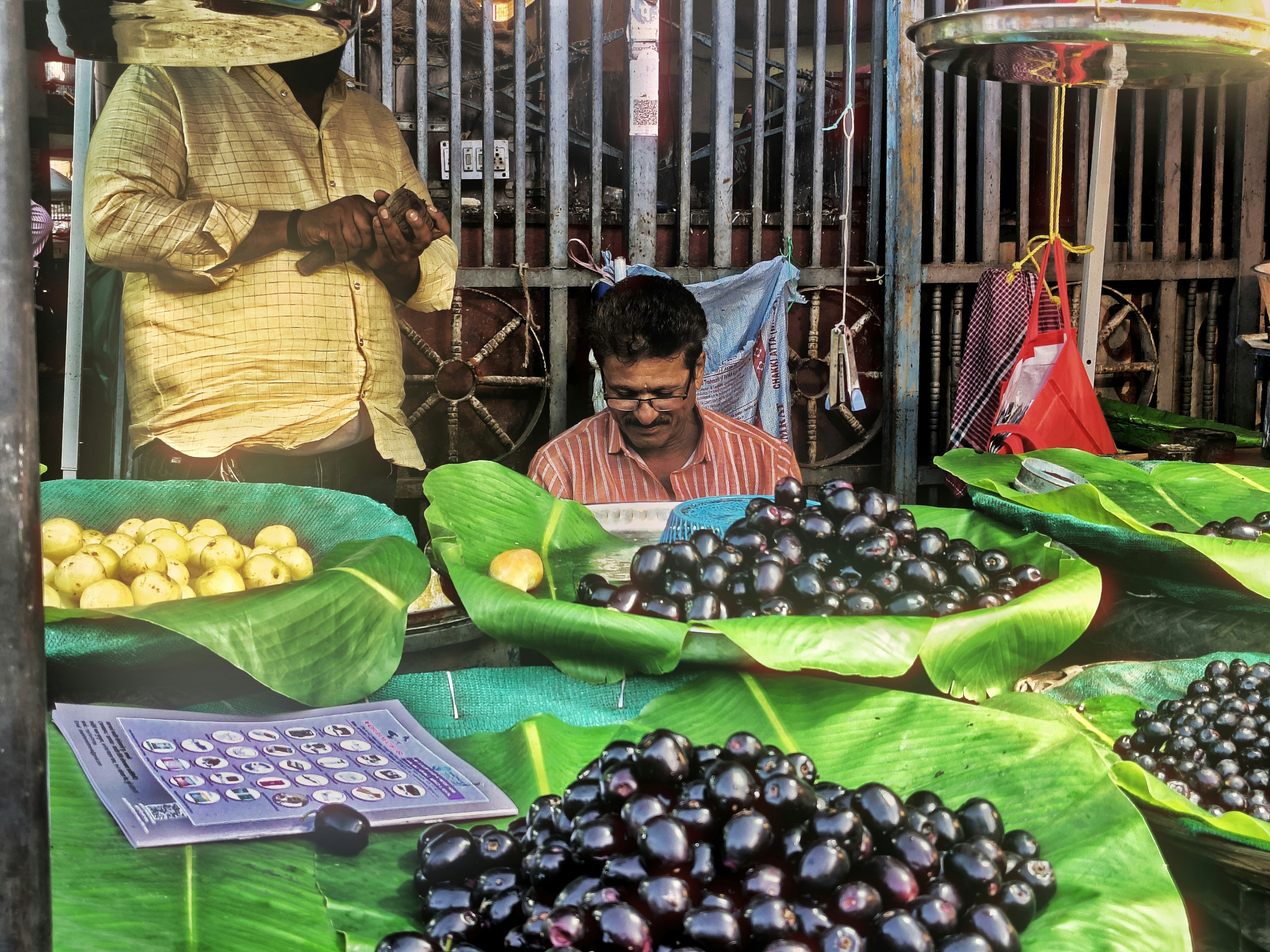 A vendor weighing dark purple jamun fruit on banana leaves at a market stall