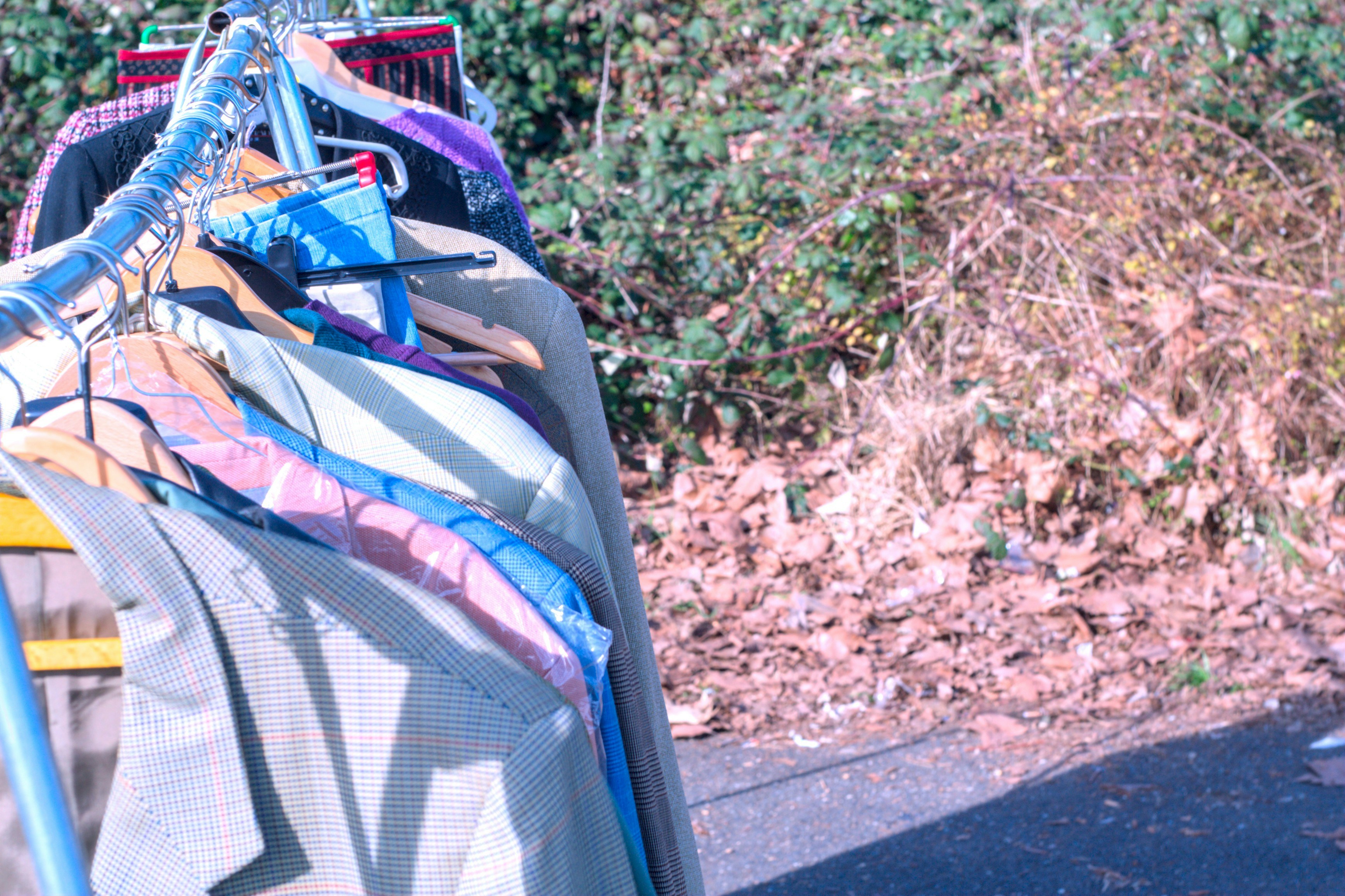 A metal clothing rack with various jackets on hangers outdoors near dry foliage