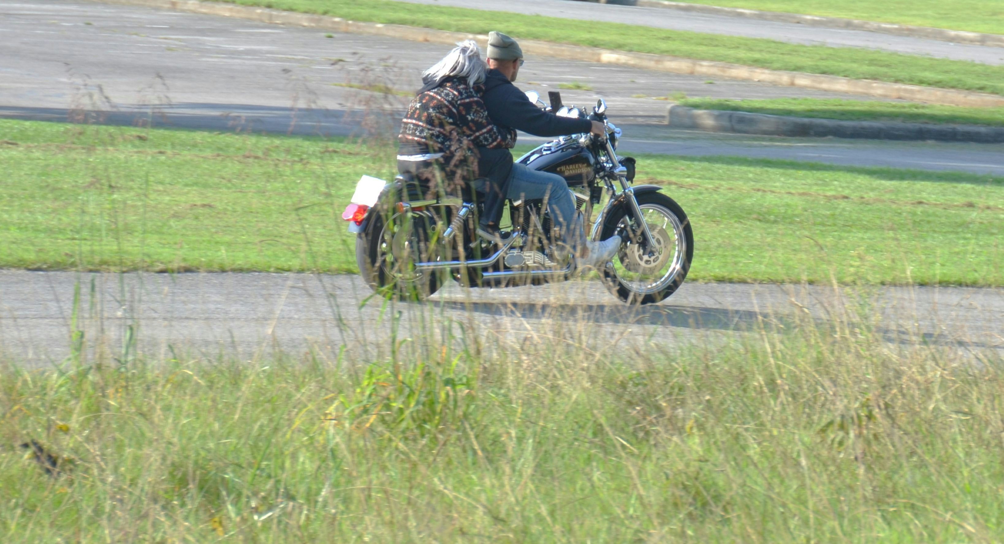 Zwei Personen fahren mit einem schwarzen Motorrad auf einer Asphaltstraße in der Nähe eines grasbewachsenen Feldes