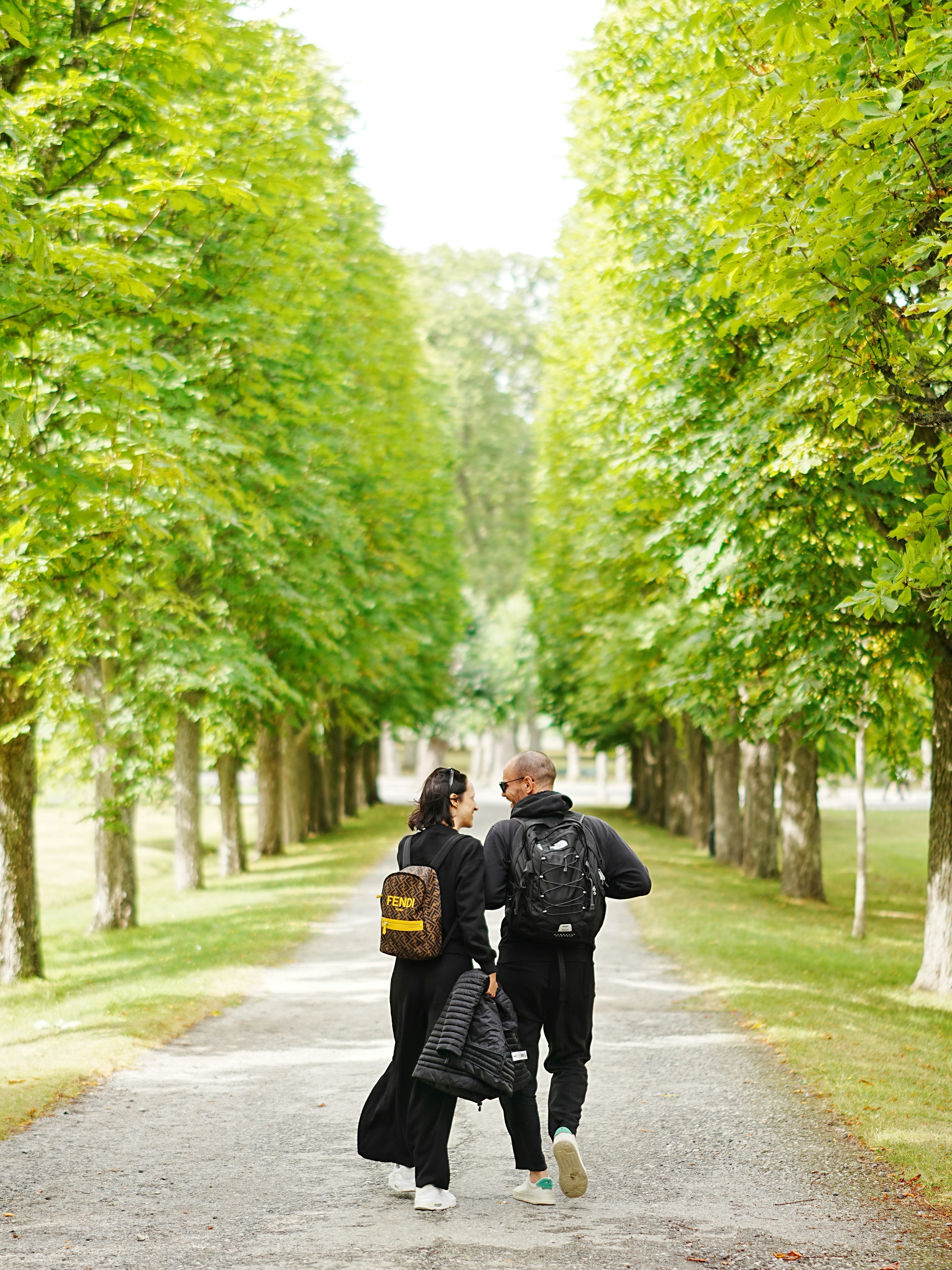 Two people with backpacks walking along a path lined with tall green trees