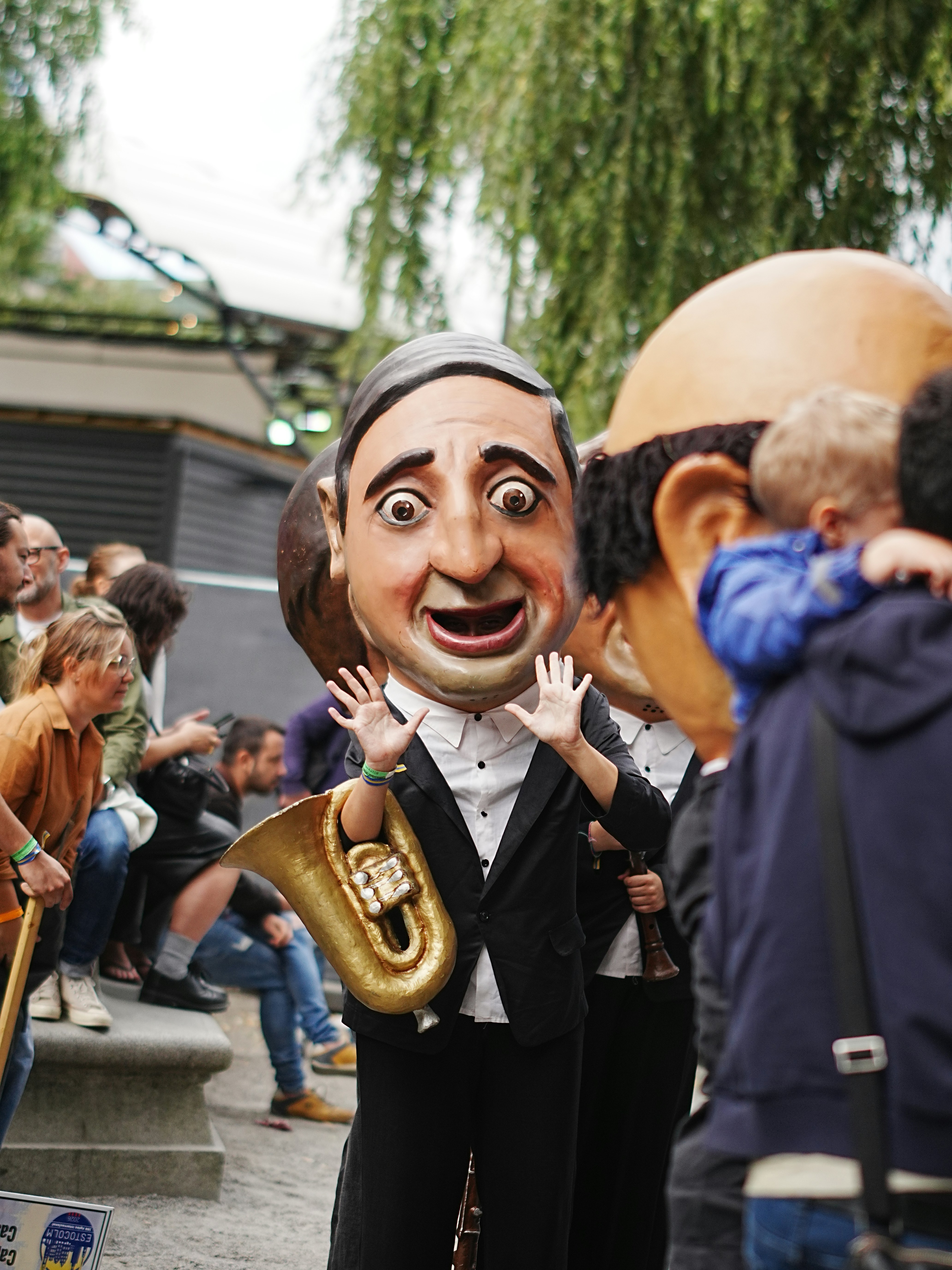 A performer in a large caricature mask and suit waving at a crowd