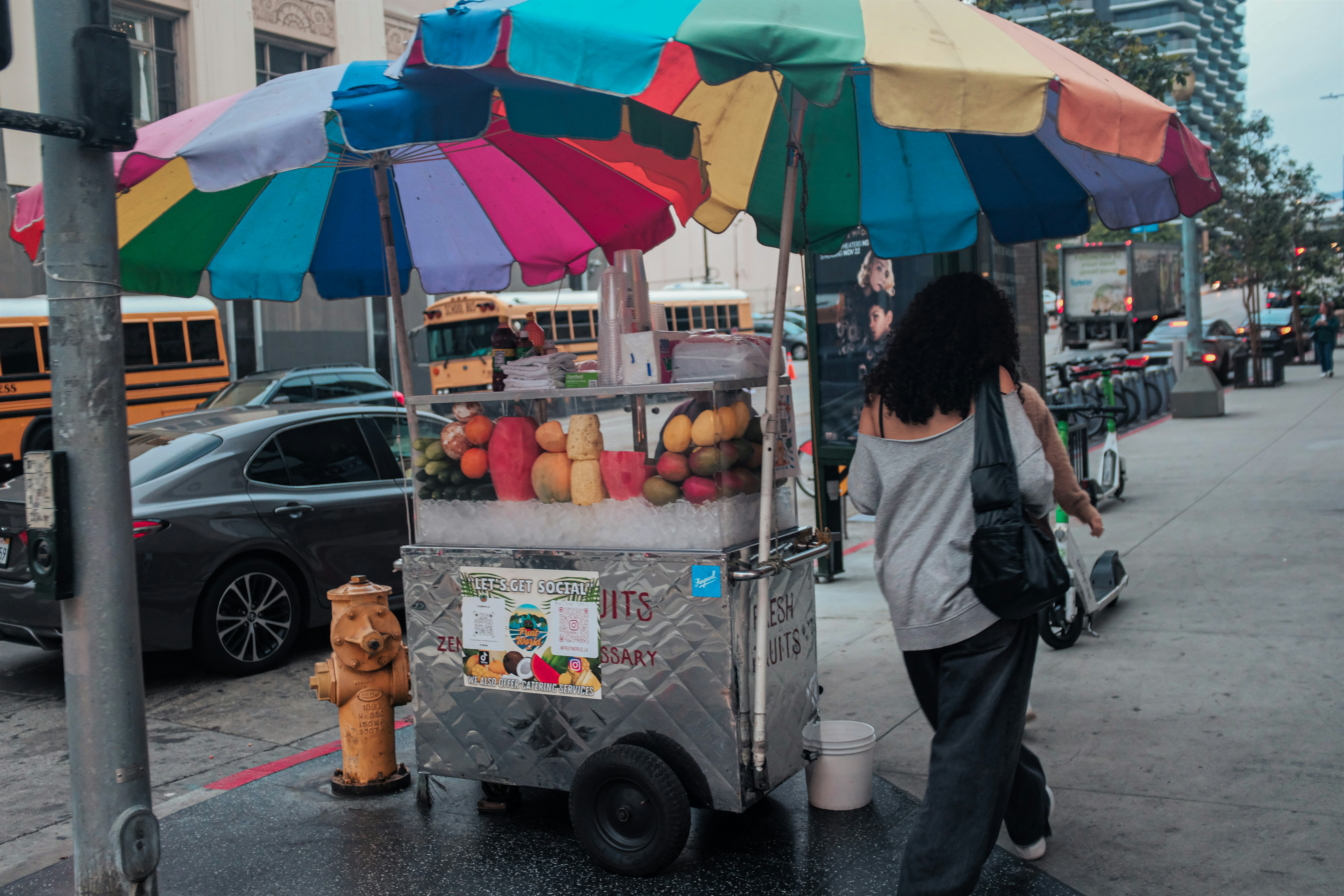 A fruit cart with a colorful umbrella on a city sidewalk near a fire hydrant