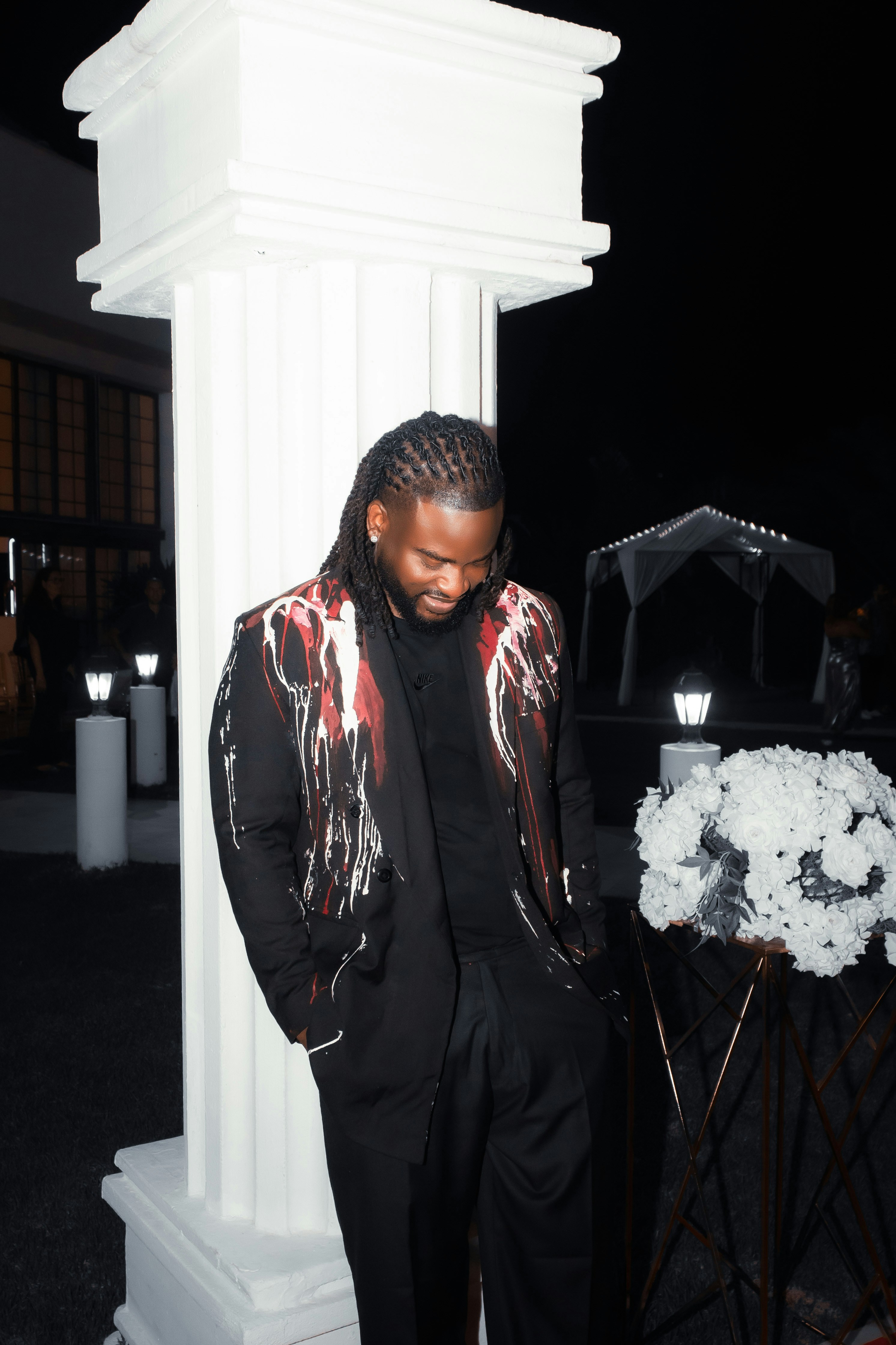 A man with braided hair wearing a black paint-splattered blazer by a white pillar