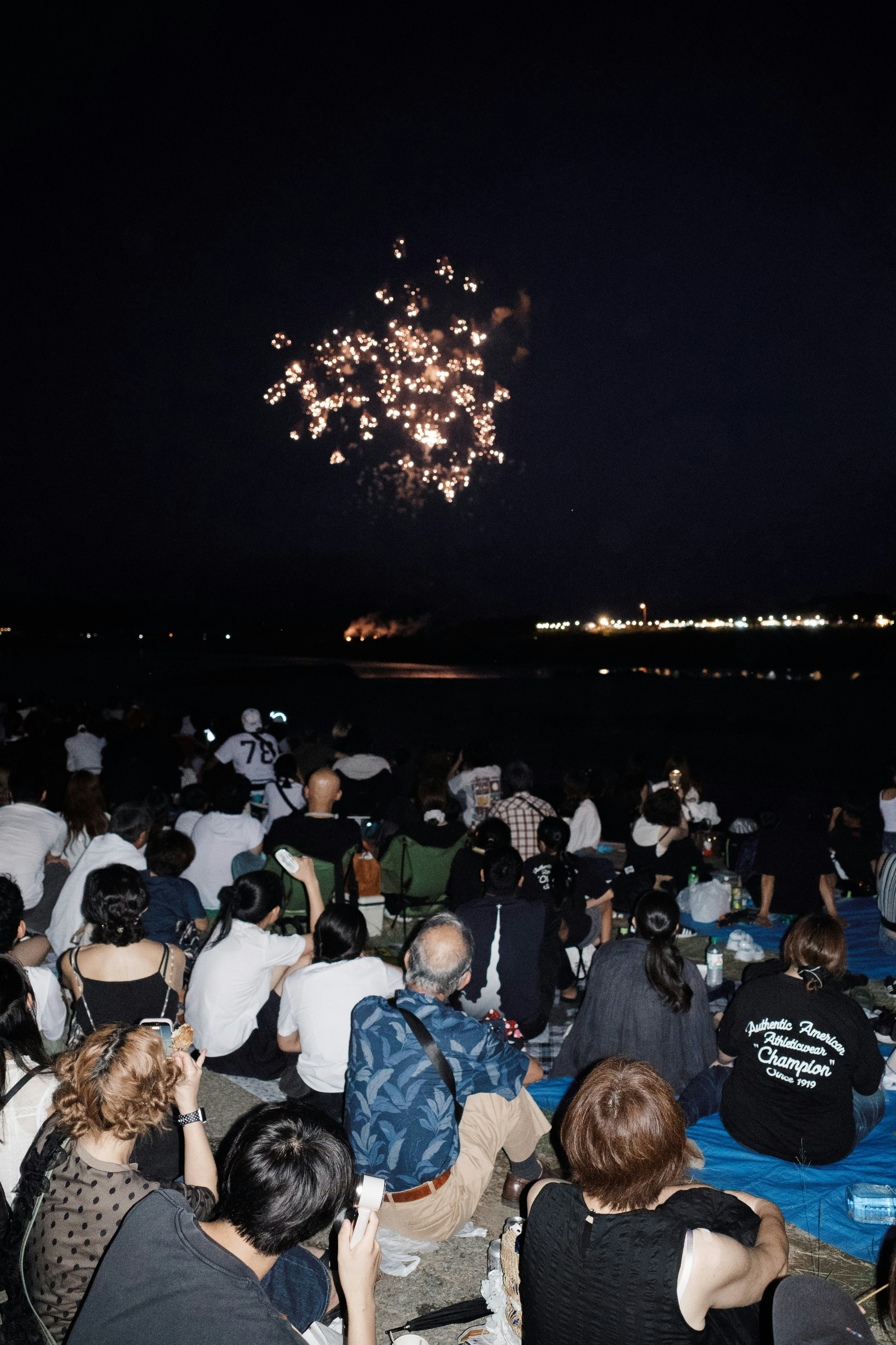 A crowd of people sitting on blue tarps watching fireworks over a dark lake