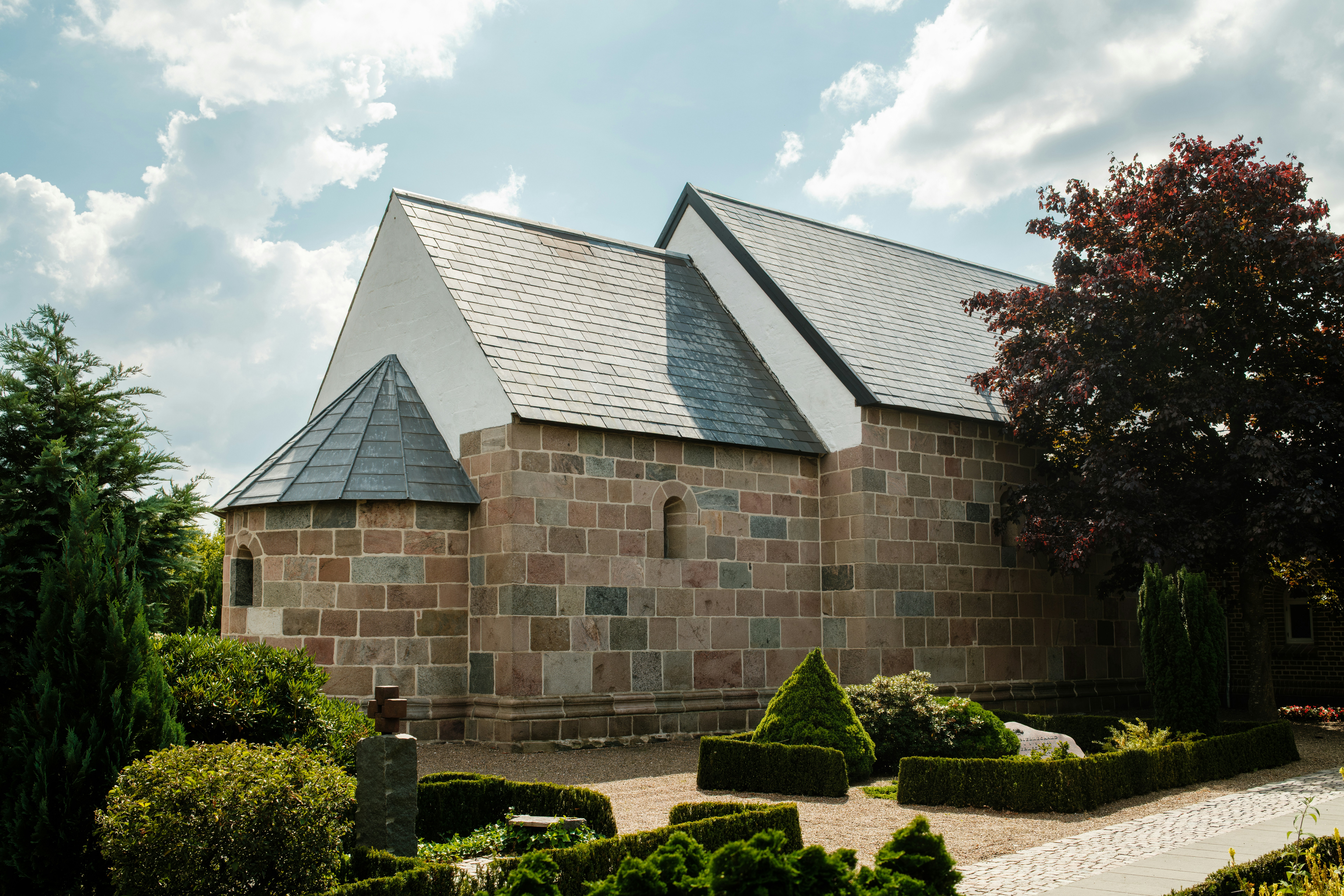 A stone church with a slate roof in a garden with manicured hedges