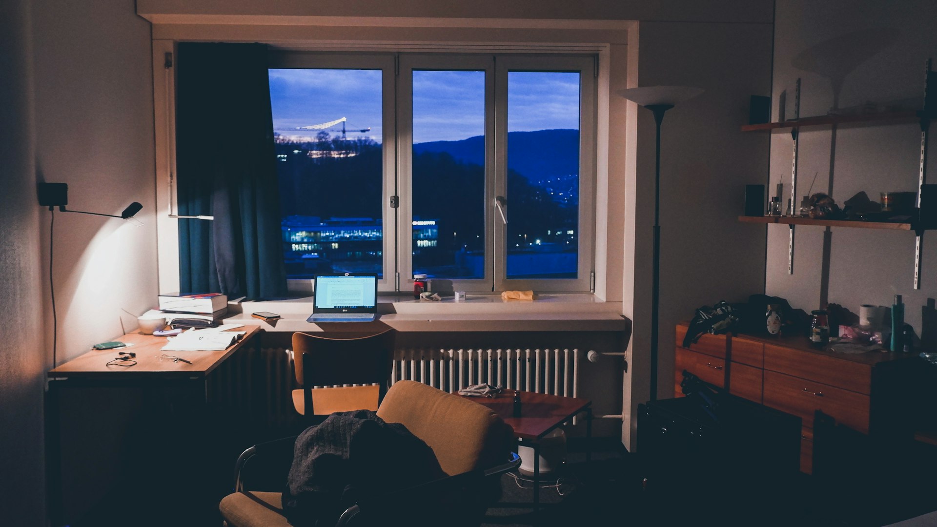 Laptop on a desk by a window at night, with a desk lamp lighting a stack of books