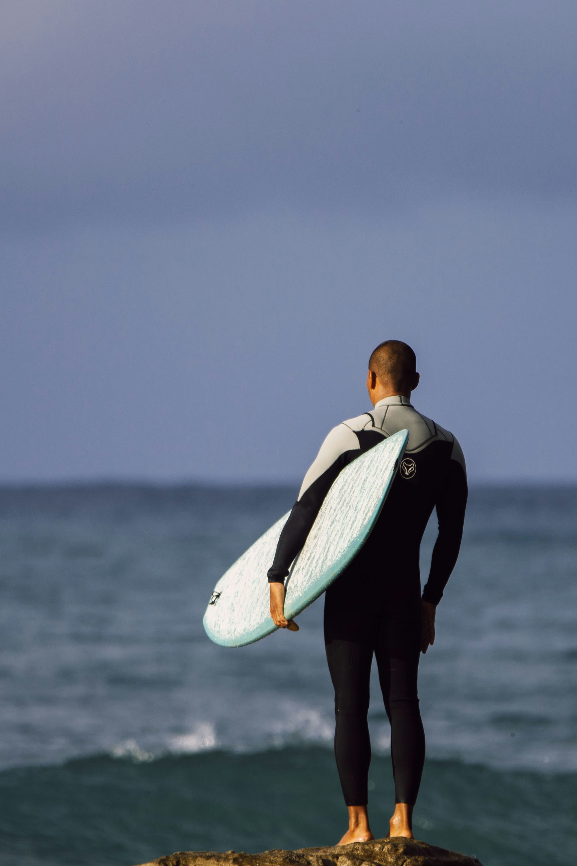 A person in a wetsuit holding a surfboard on a rock by the ocean