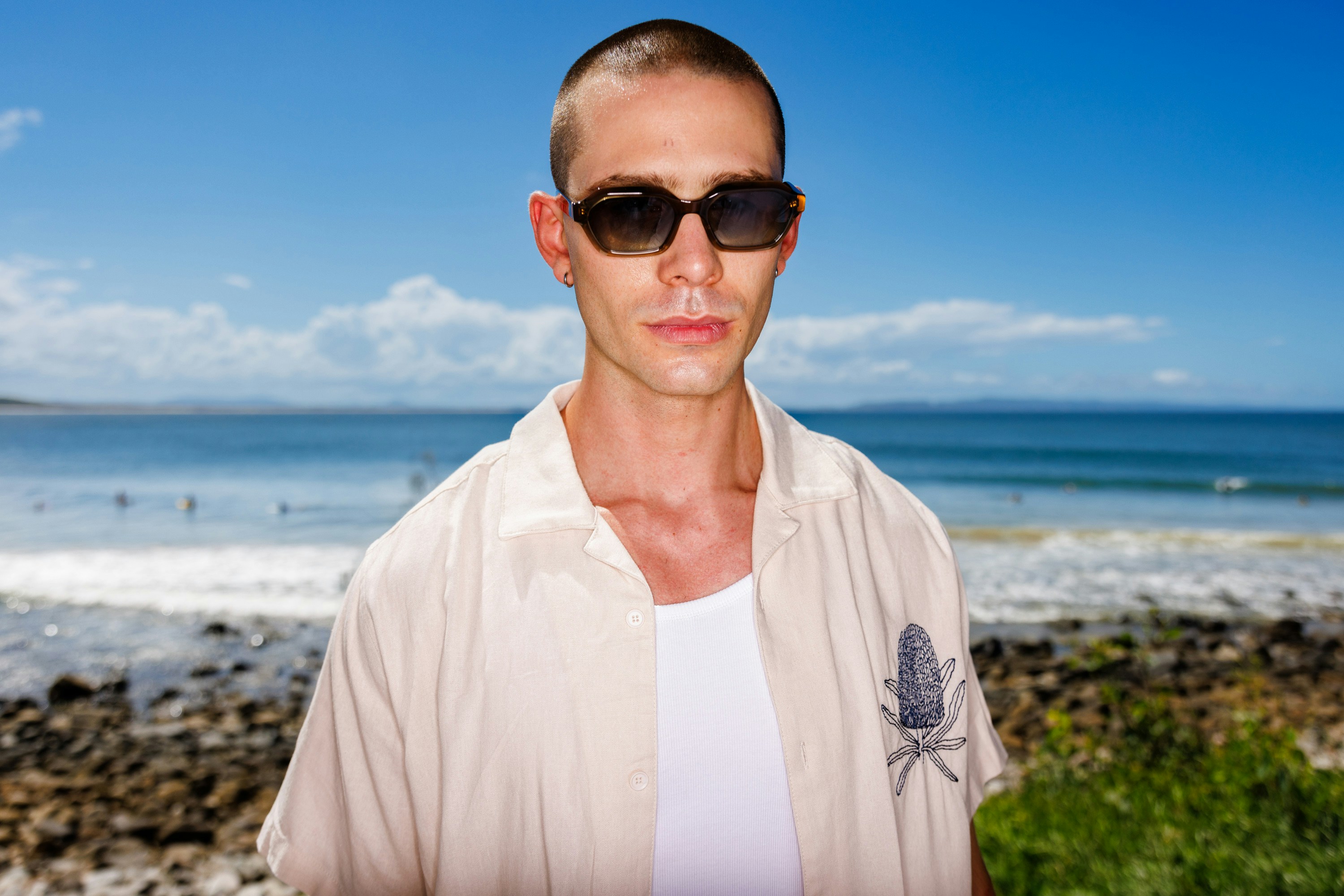 A man with a buzz cut wearing sunglasses and a beige shirt by the ocean