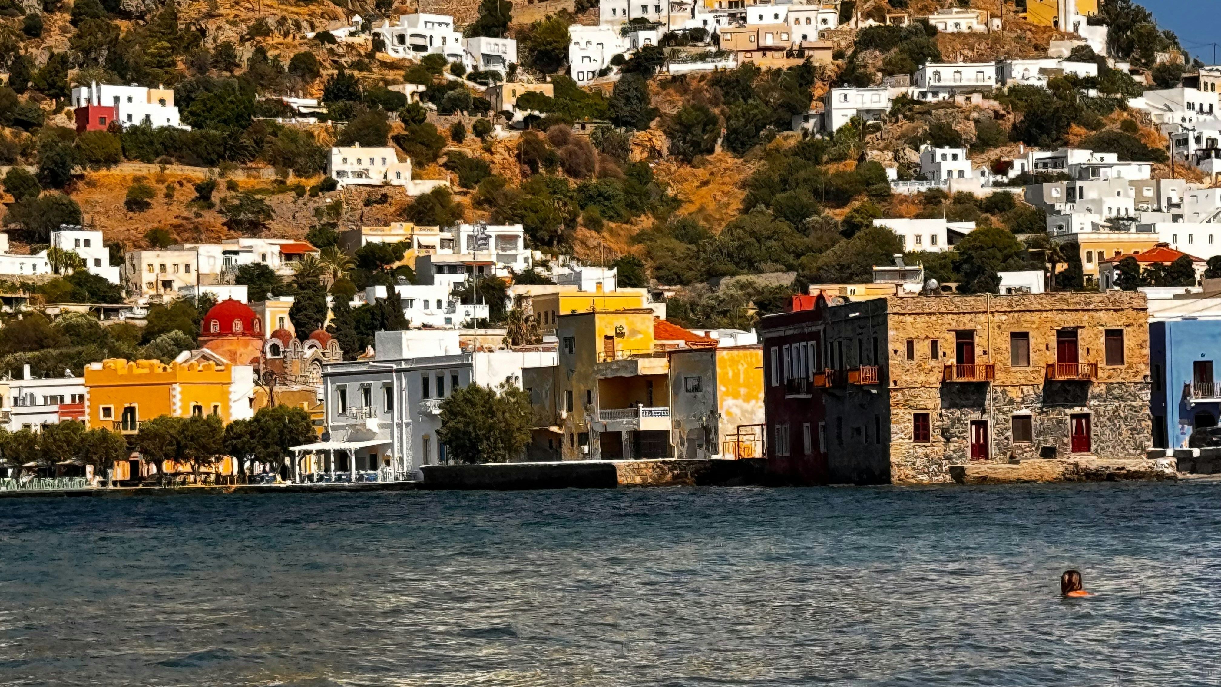 A person swimming in the sea before colorful buildings on a hillside in Symi