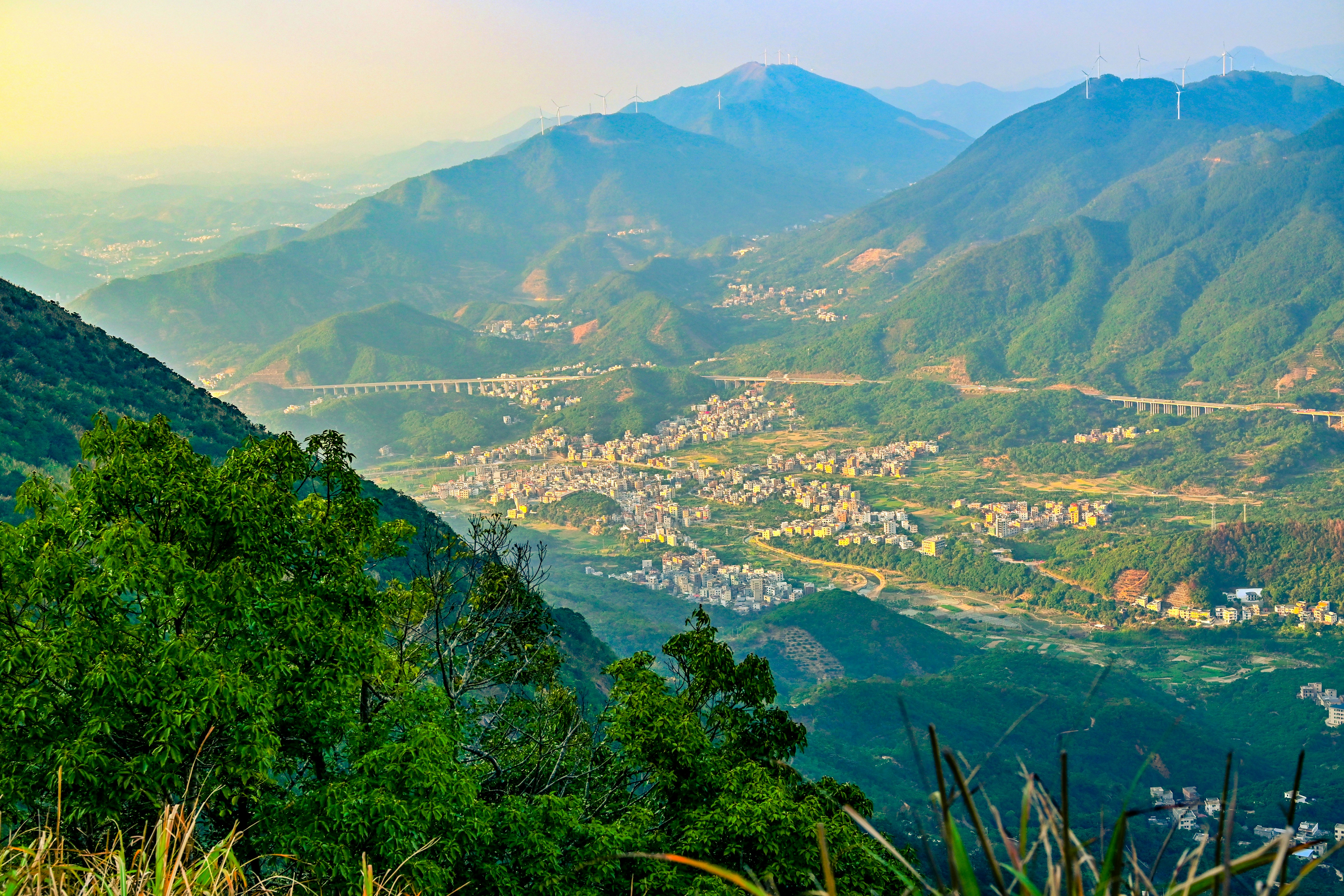 A town nestled in a green mountain valley with a long bridge and wind turbines