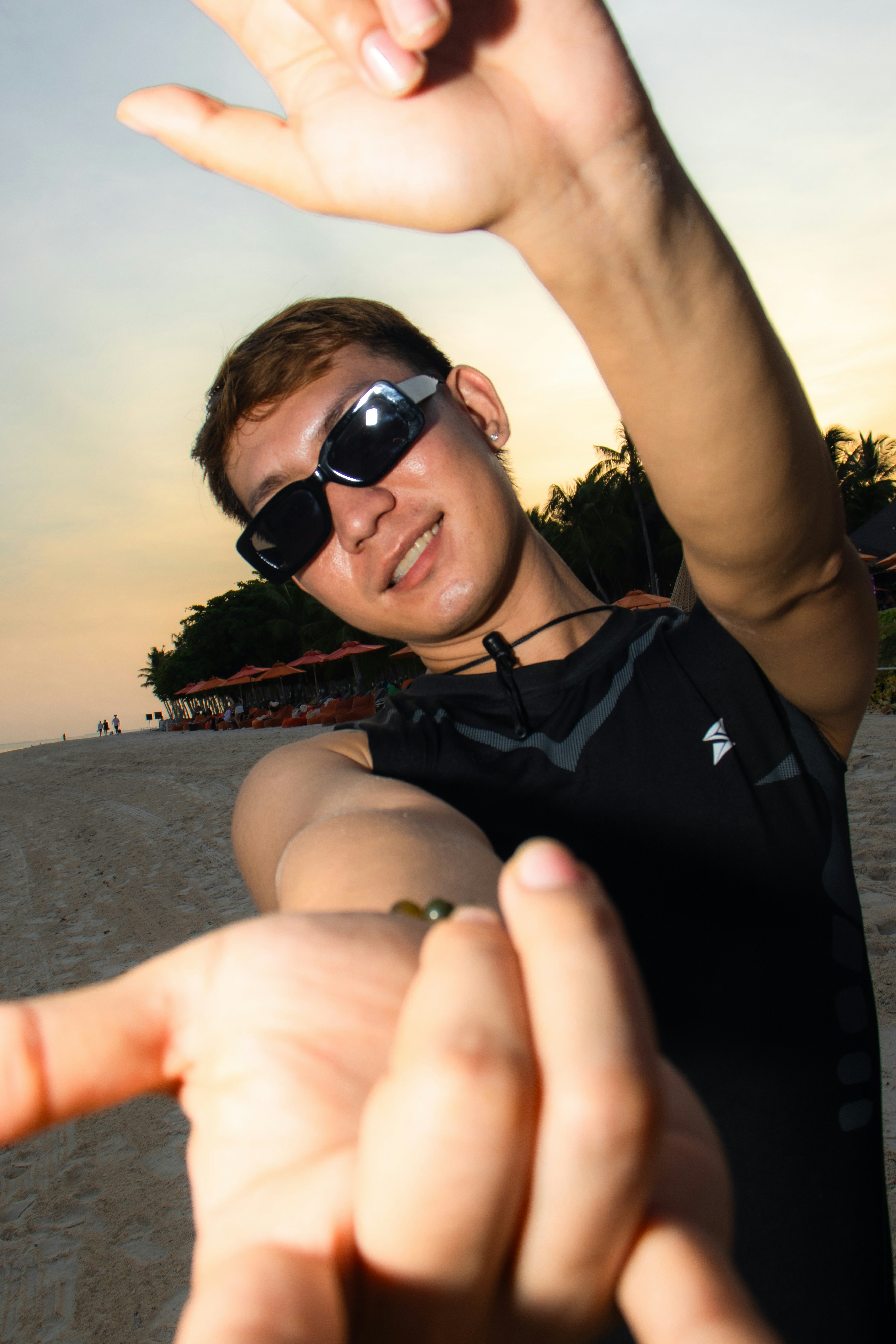 A man in sunglasses reaching toward the camera on a sandy beach at sunset