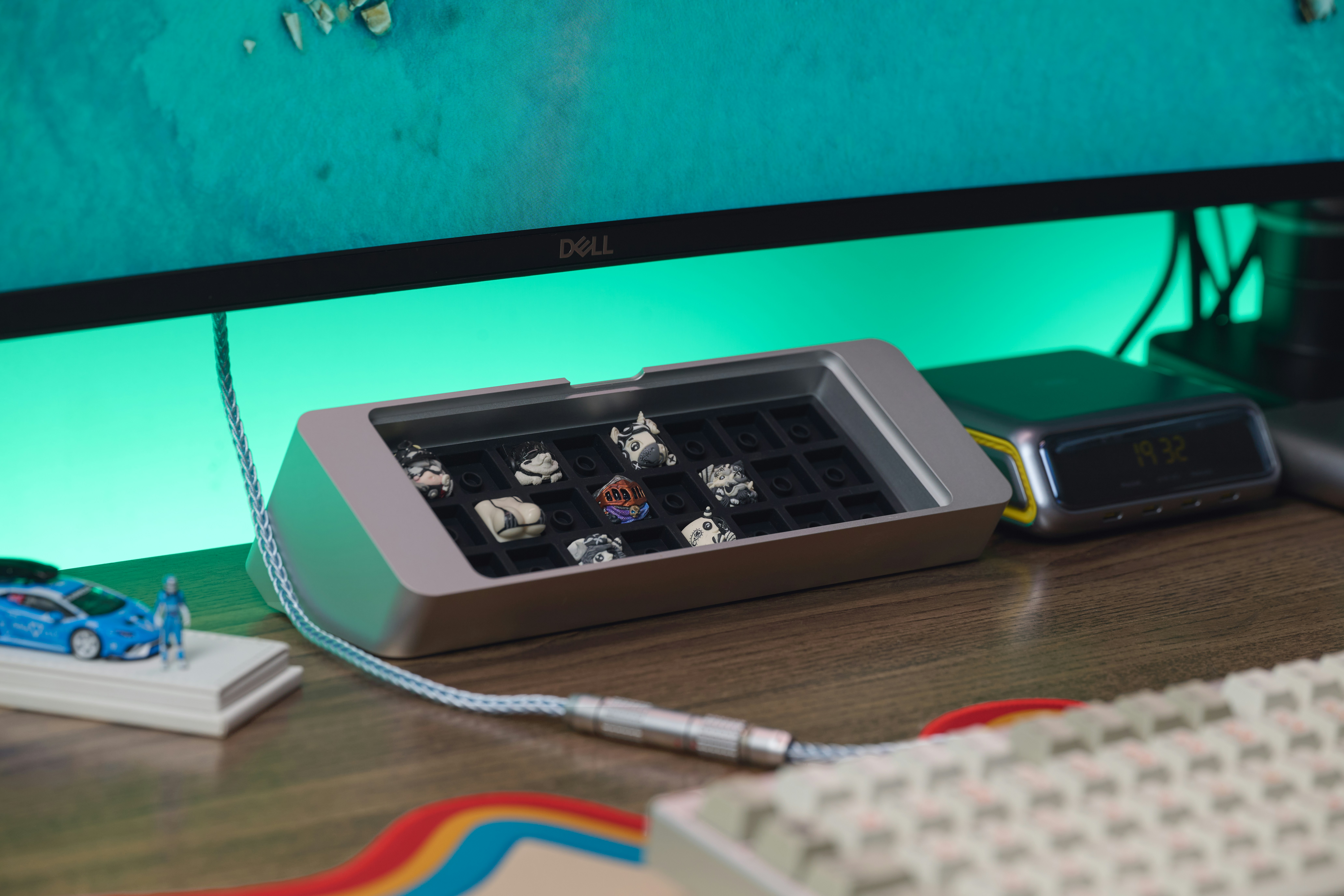 A silver artisan keycap display case on a wooden desk with a monitor