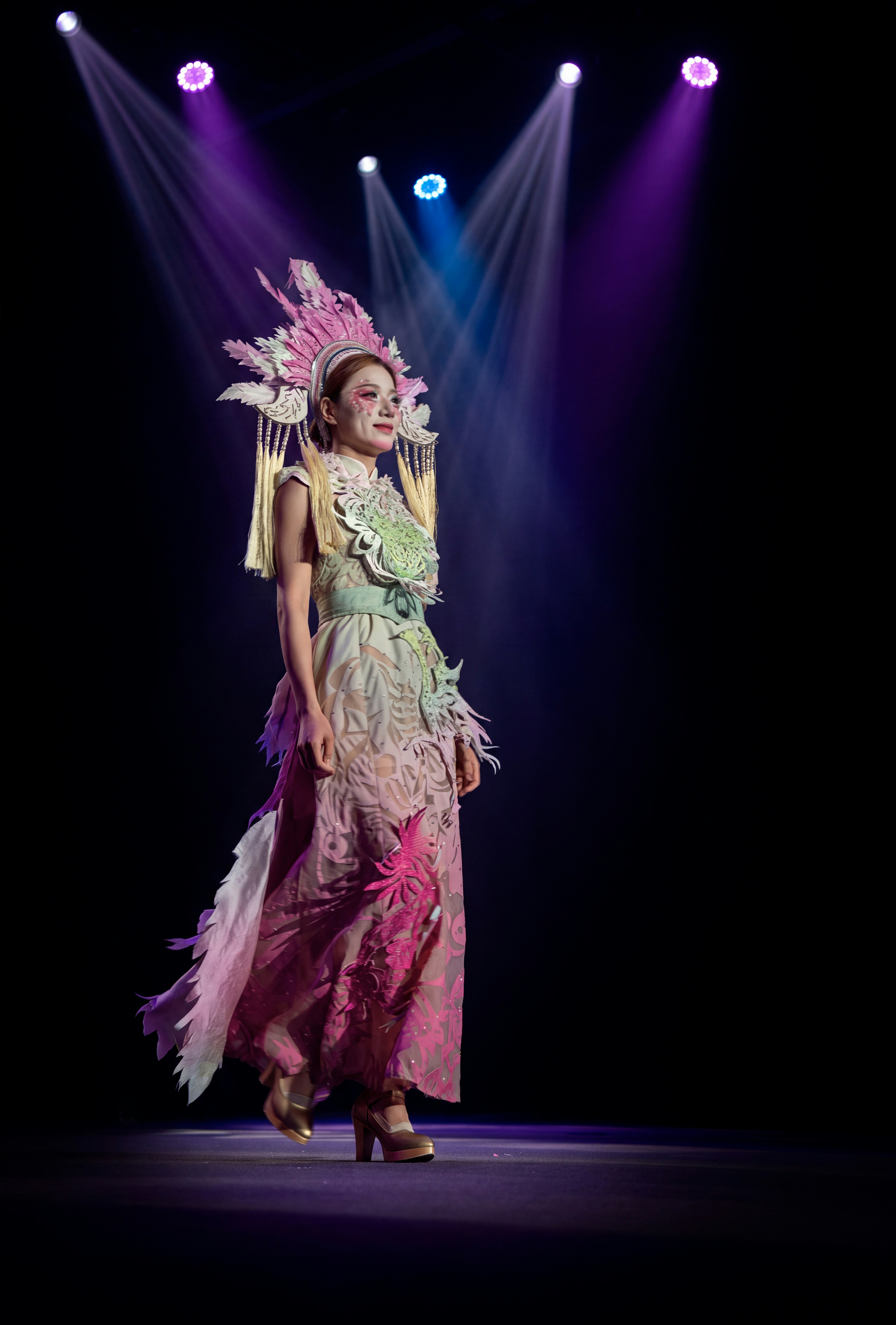 A woman in a pink feathered headdress and patterned gown walking on a stage