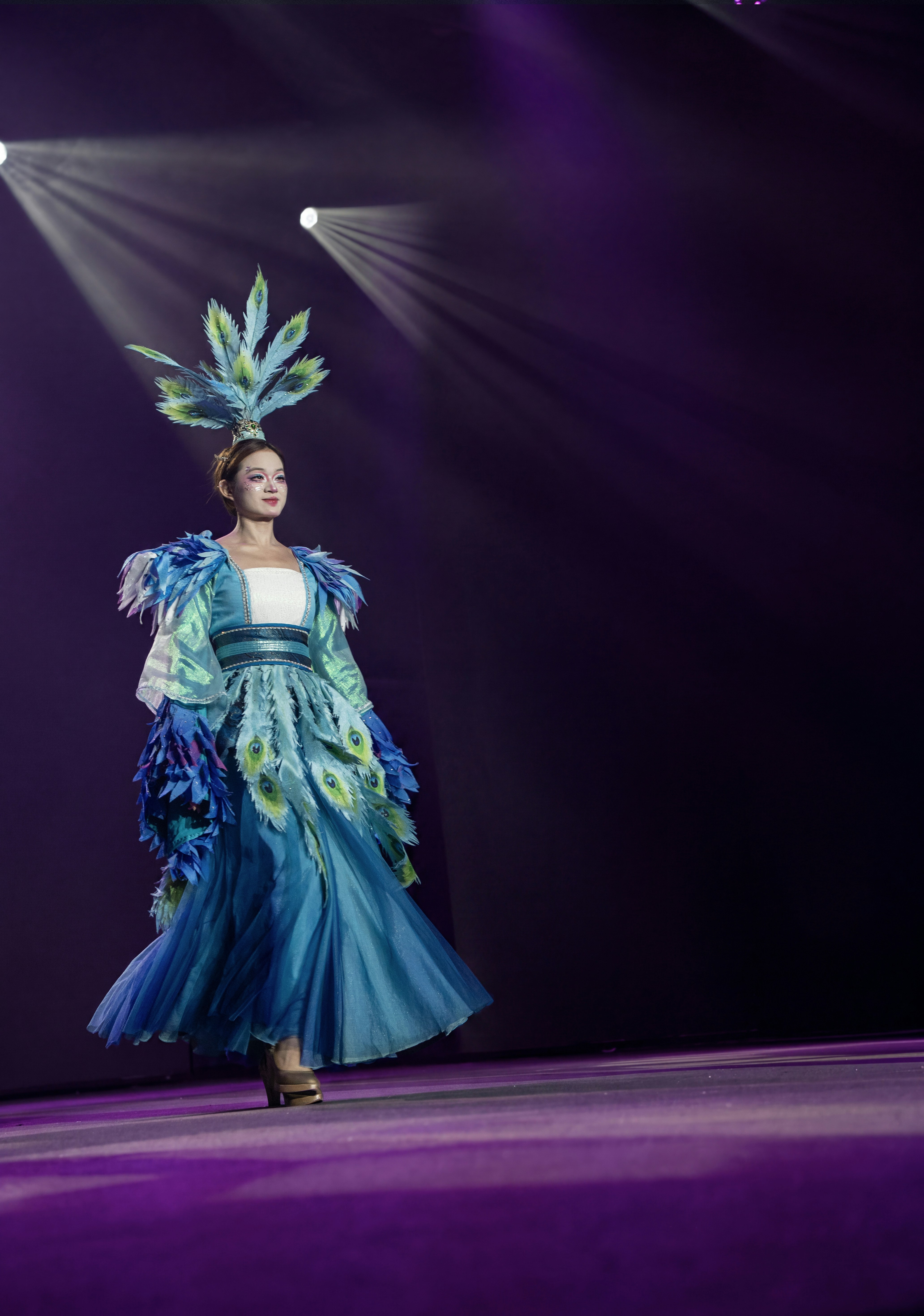 A person in a blue peacock-inspired costume walking on a stage under spotlights