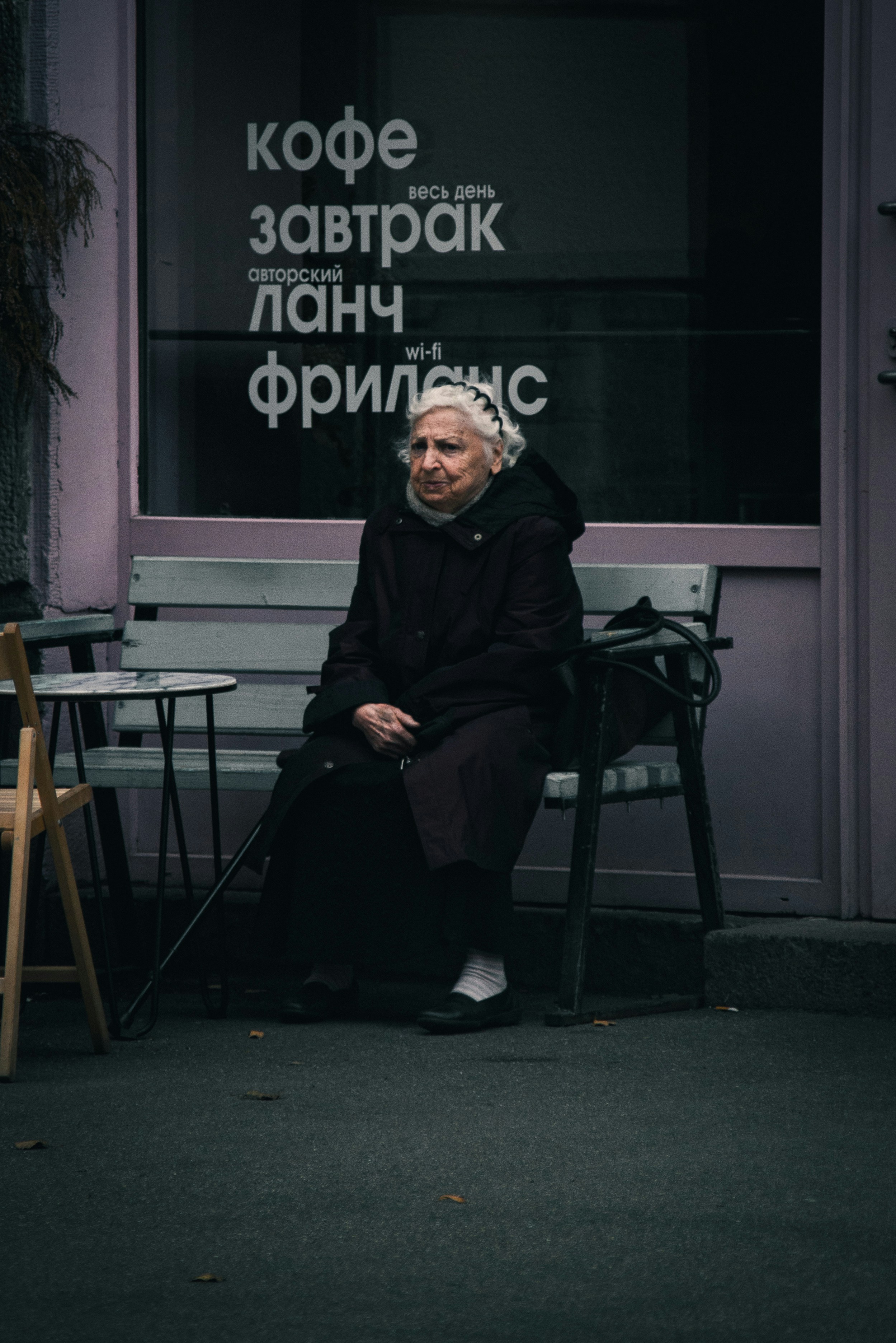 An elderly woman in a dark coat sitting on a bench outside a cafe