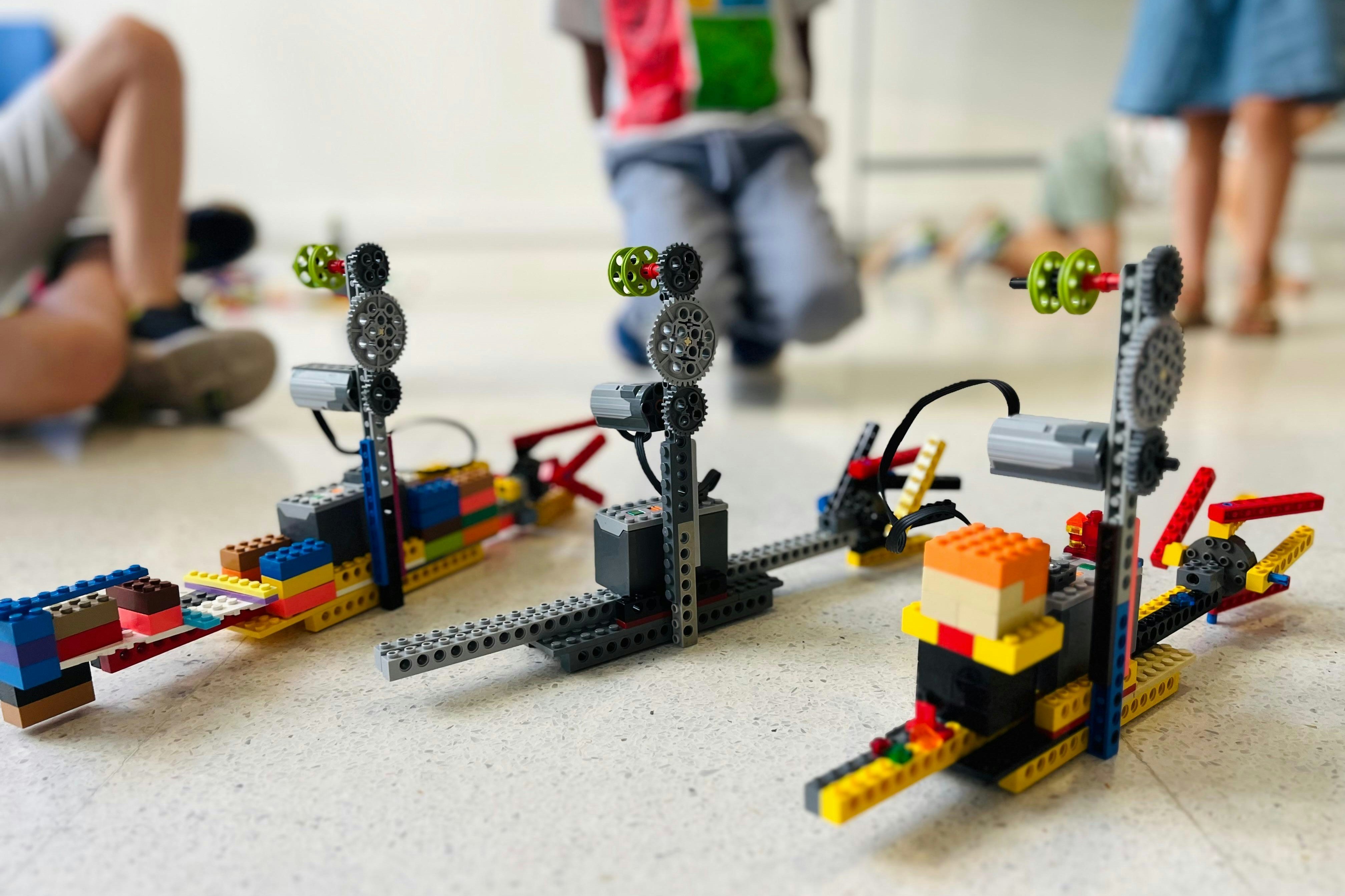 Three robotic models built from plastic bricks on a light floor with children nearby