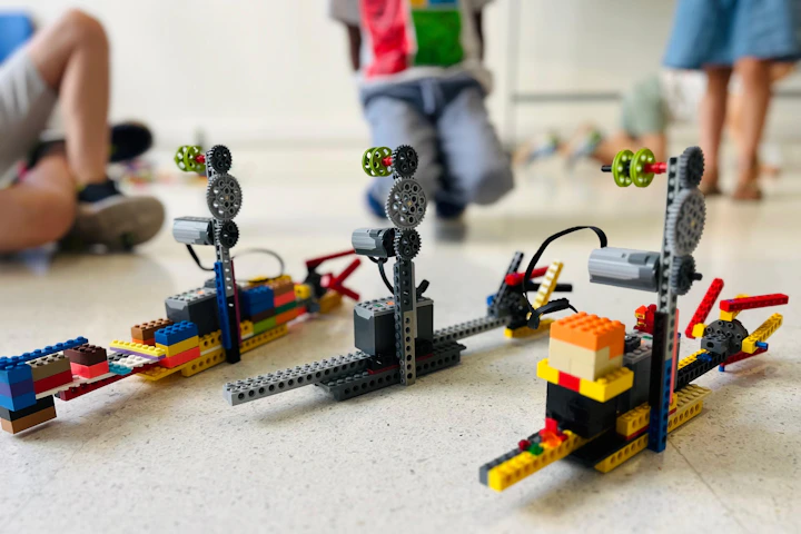 Three robotic models built from plastic bricks on a light floor with children nearby
