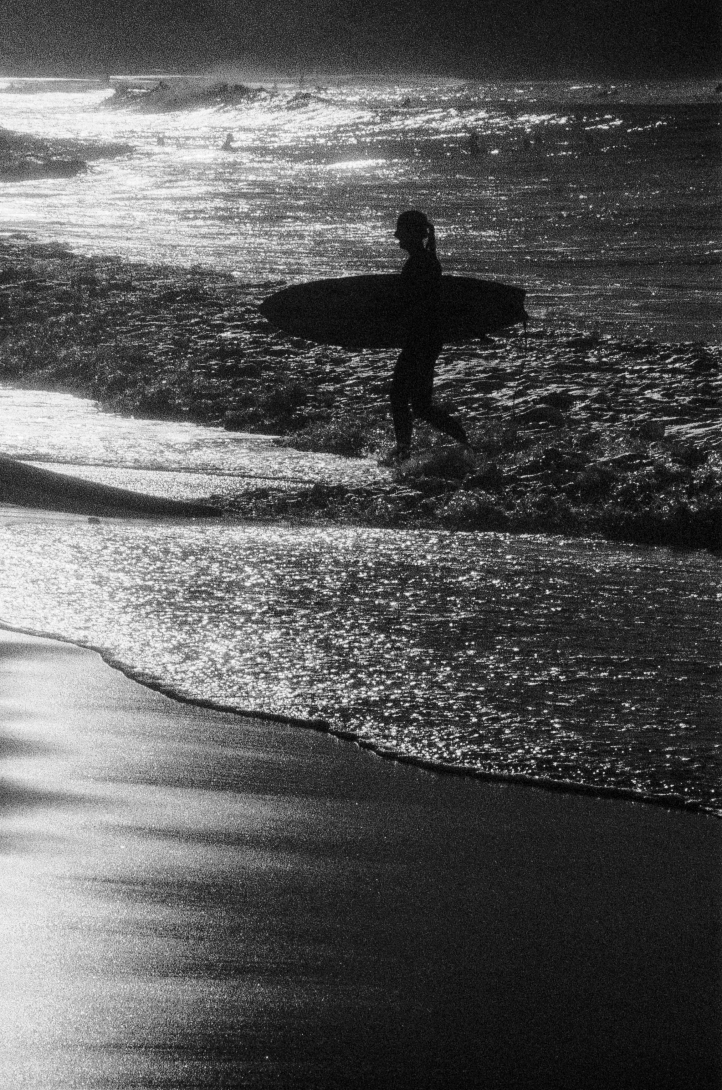 A person in silhouette carrying a surfboard through shallow water at the beach