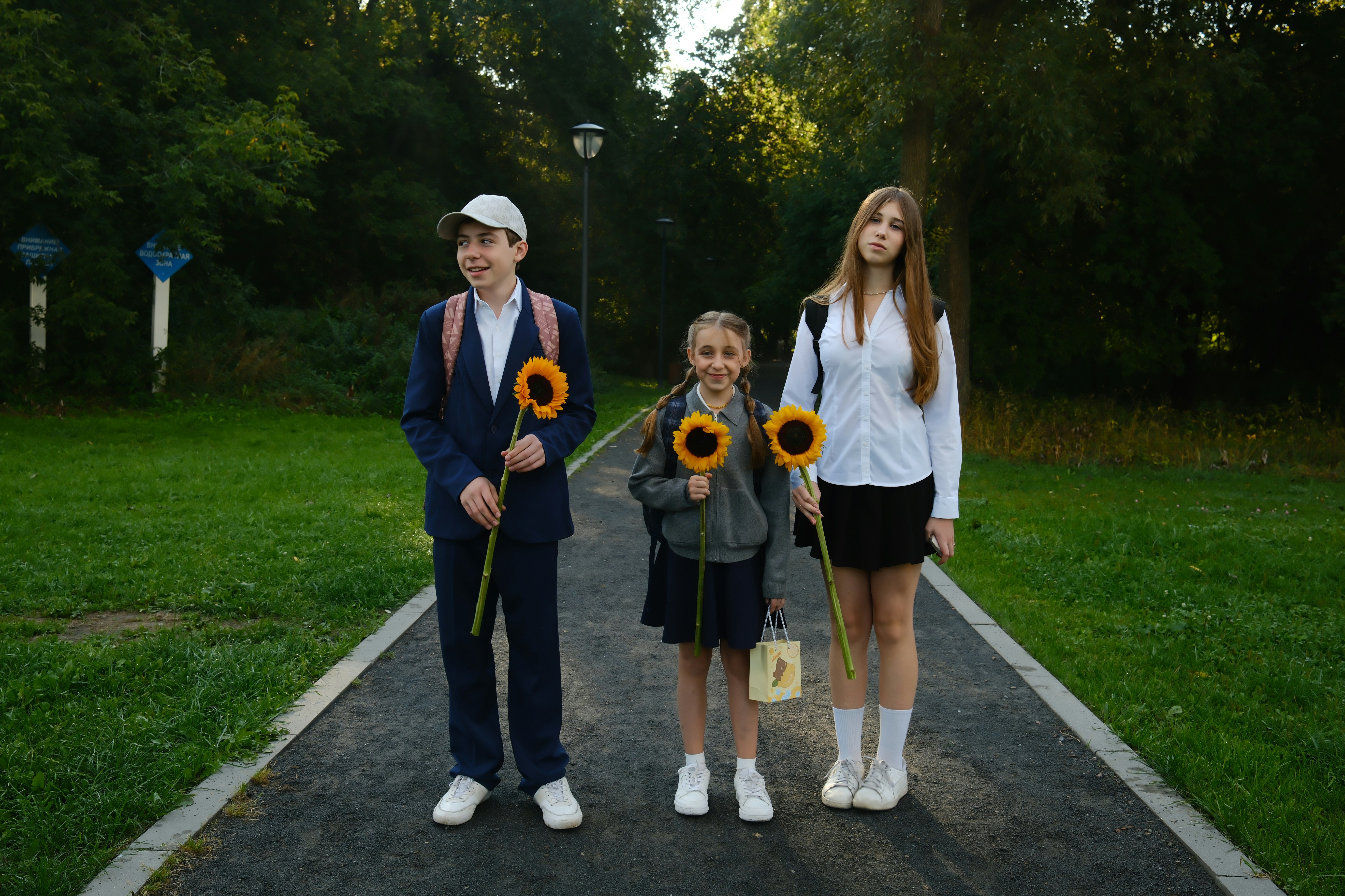 Three students holding sunflowers on a park path during the first day of school