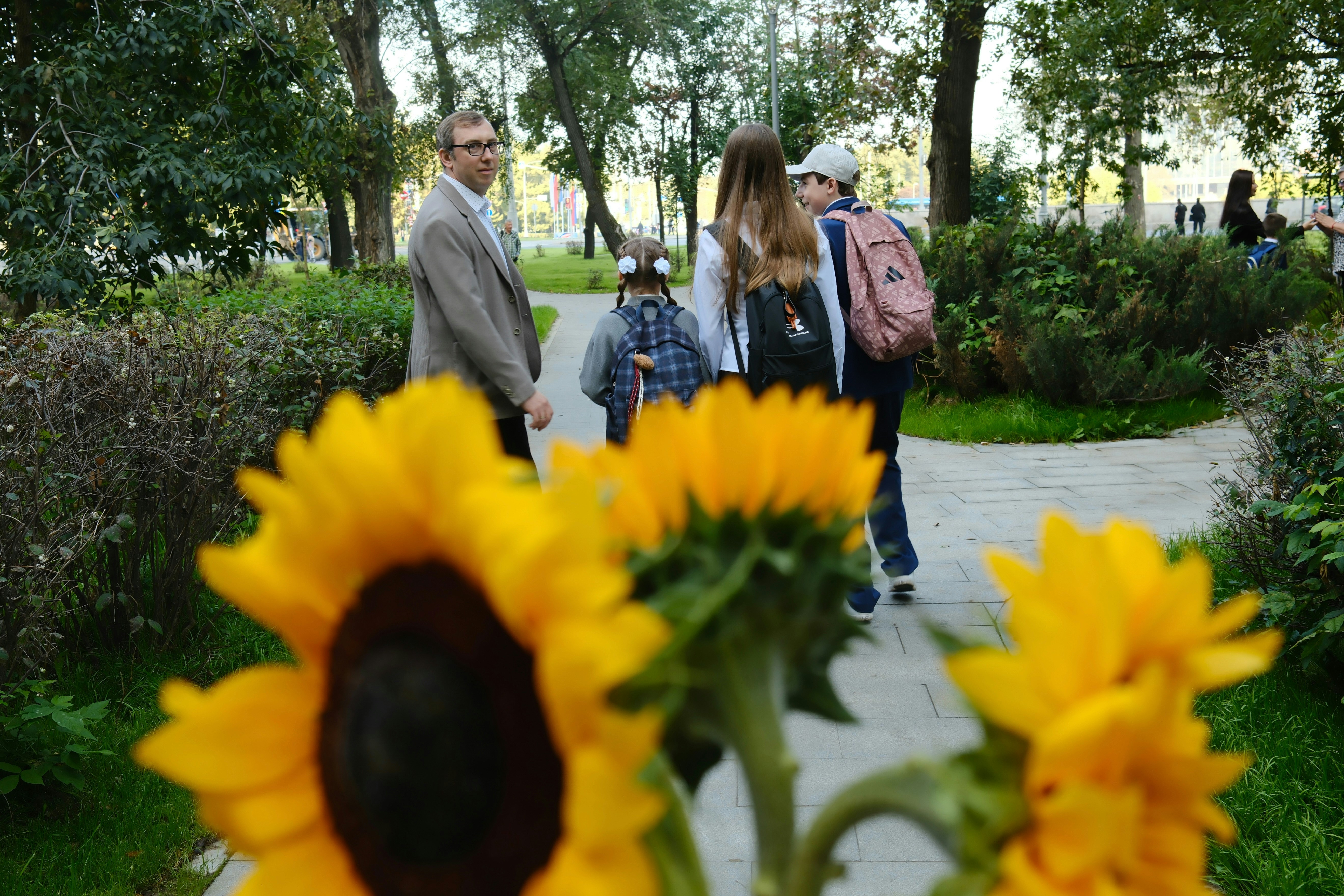A man and three children with backpacks walking along a park path near sunflowers