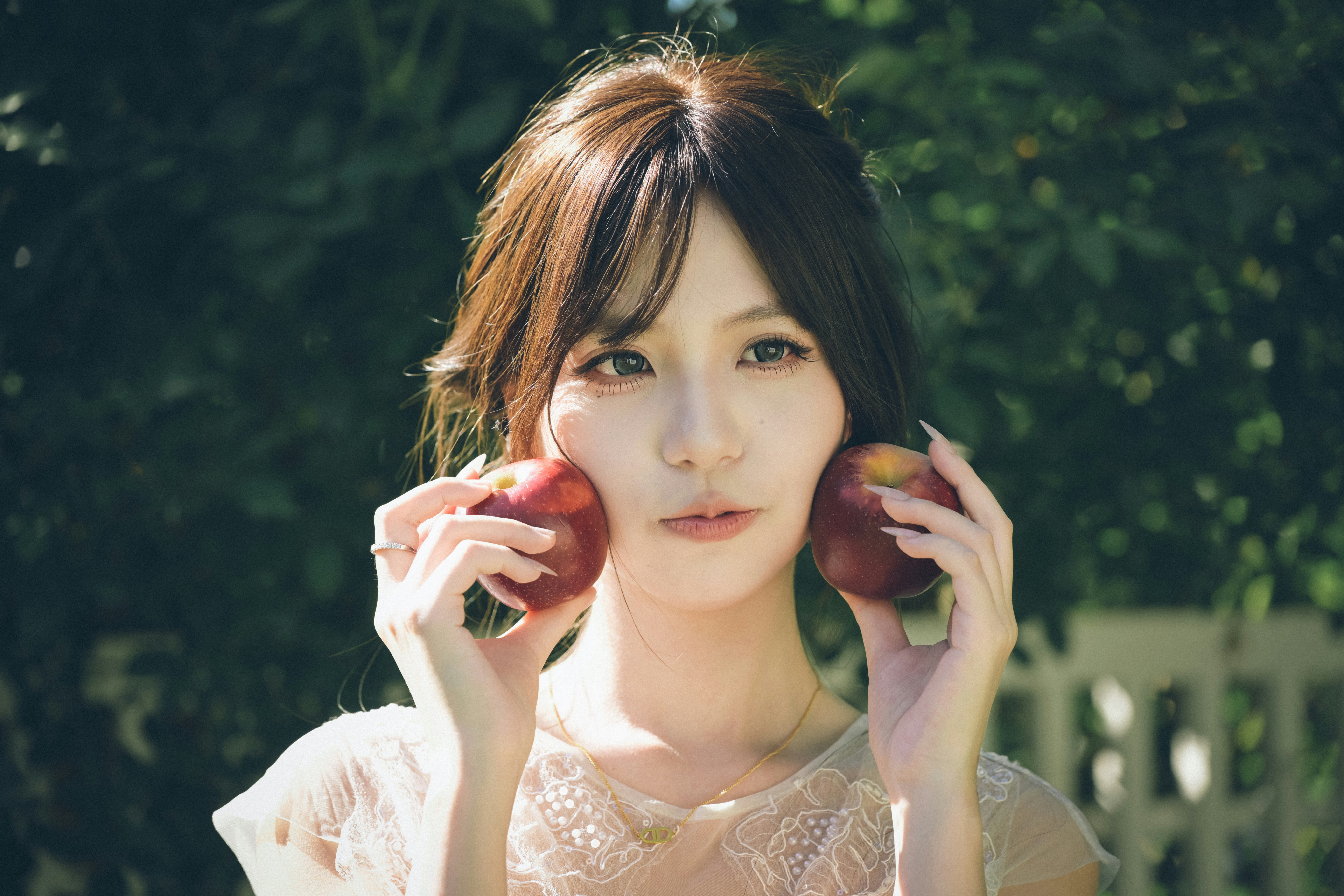 A woman holding two red apples against her cheeks in a sunlit garden