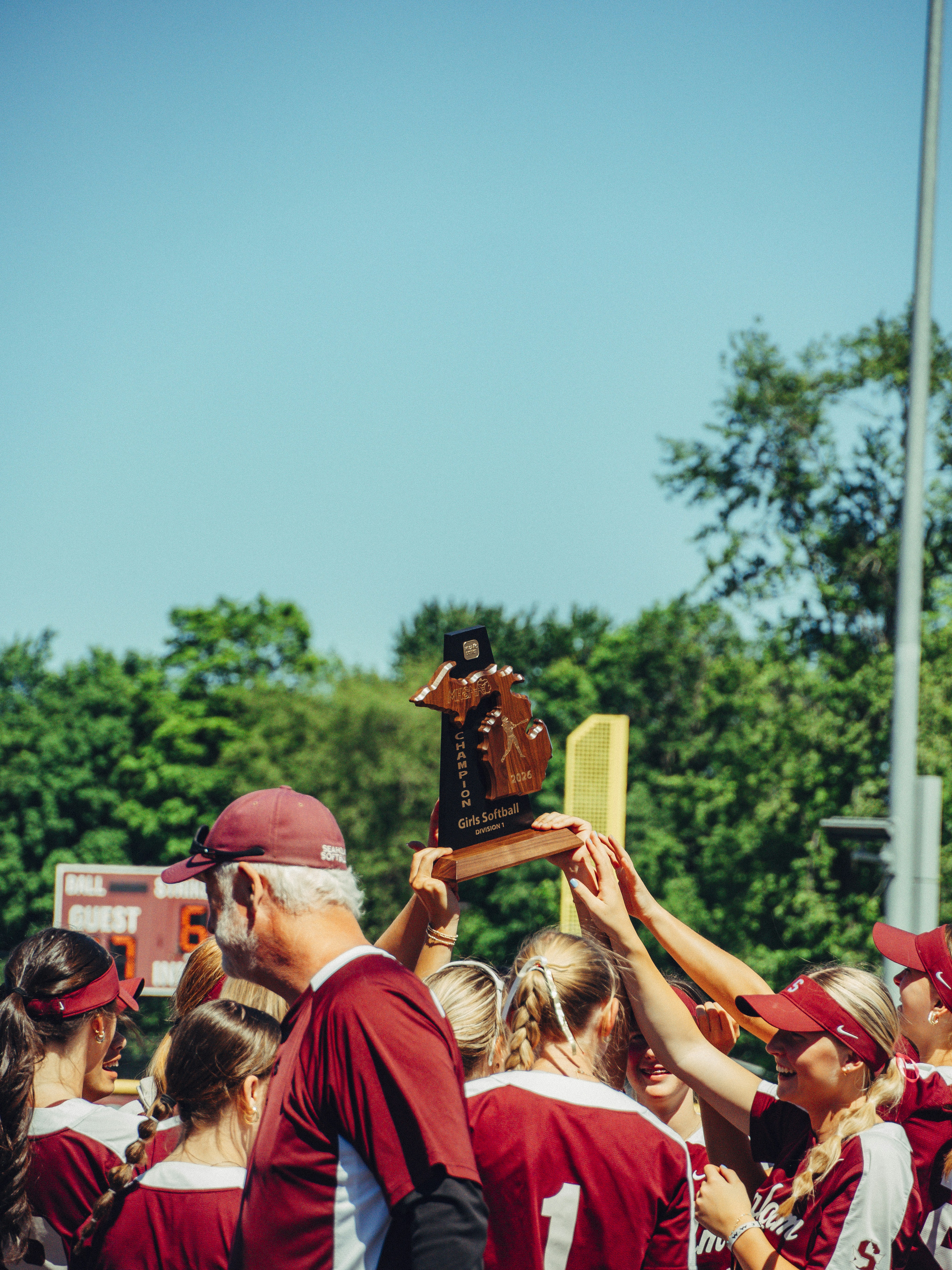 A group of softball players and a coach holding a championship trophy aloft