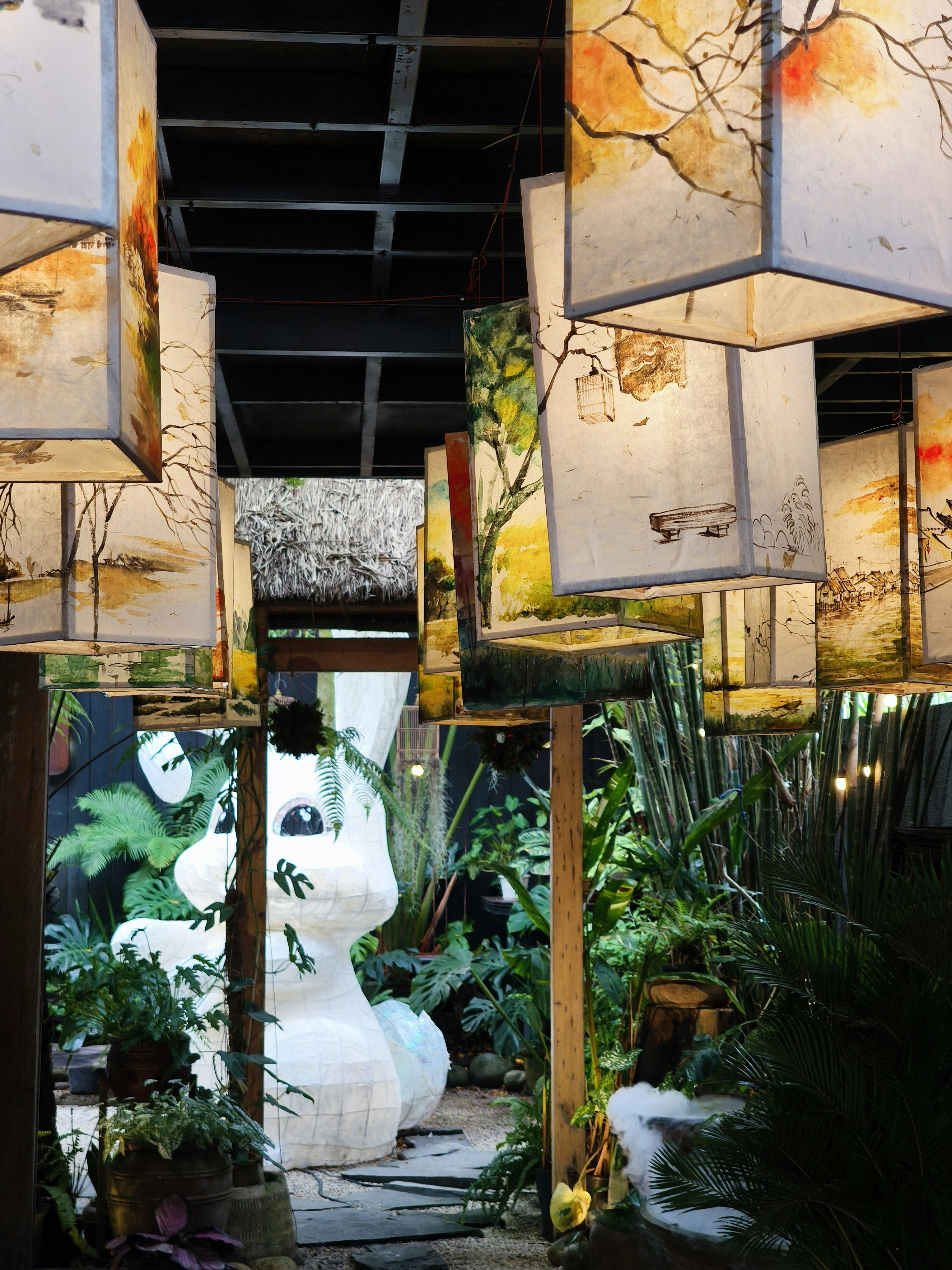 Hanging paper lanterns with painted landscapes above a garden with a large white rabbit sculpture