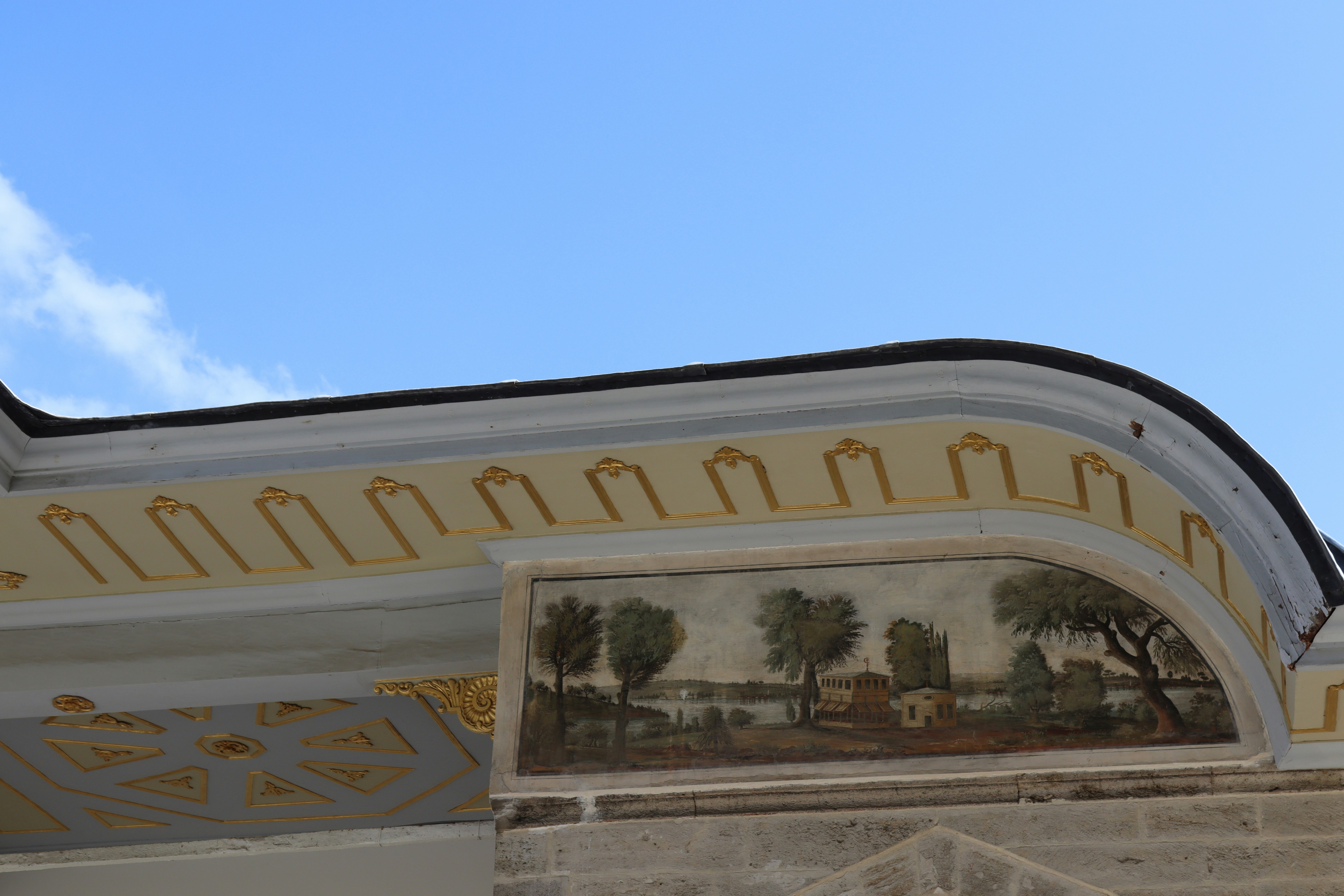A painted landscape mural on the curved cornice of a building under blue sky