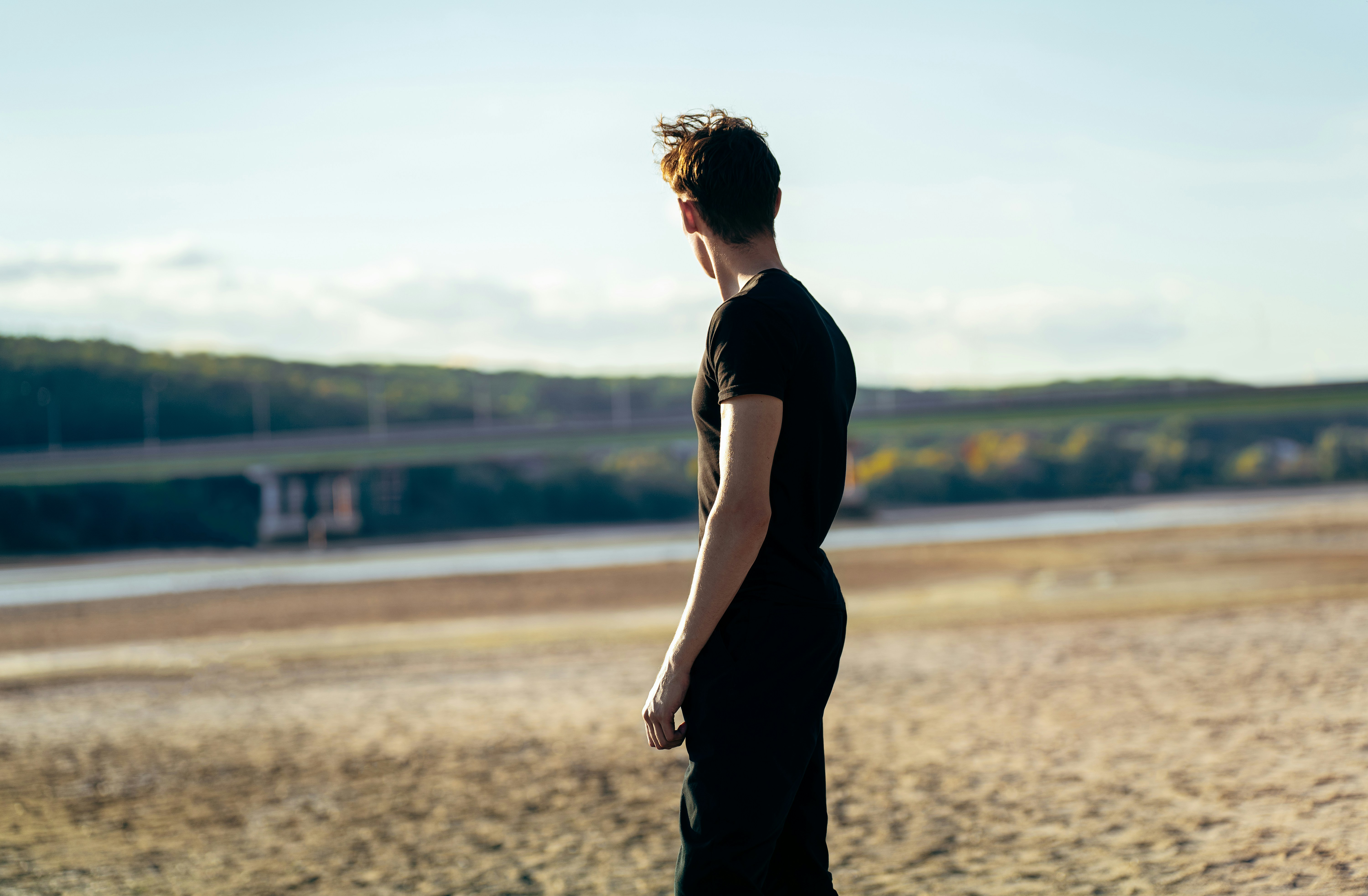 A person in a black t-shirt looking toward a distant bridge over water