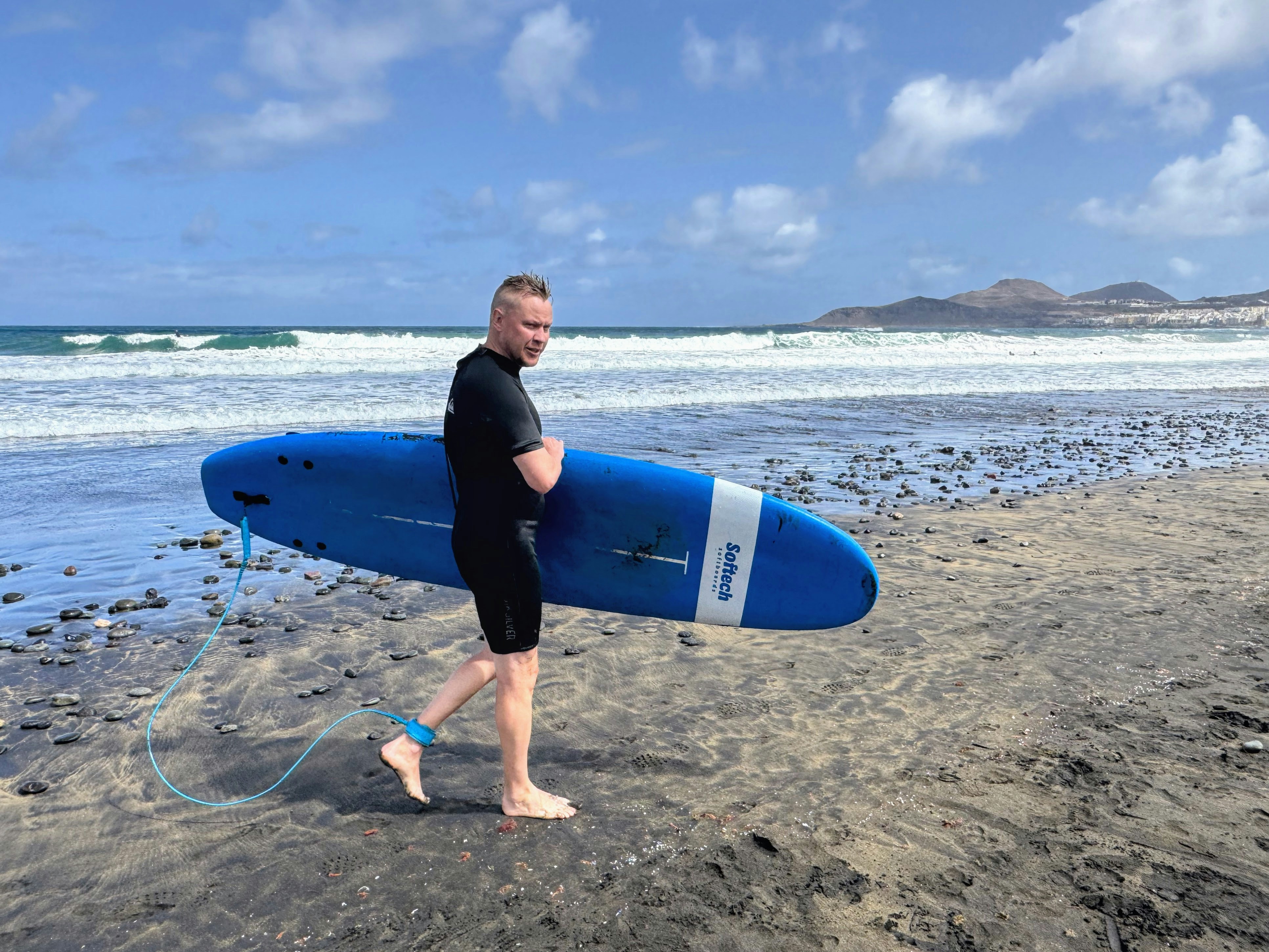 A man in a black wetsuit carrying a blue surfboard on a dark beach