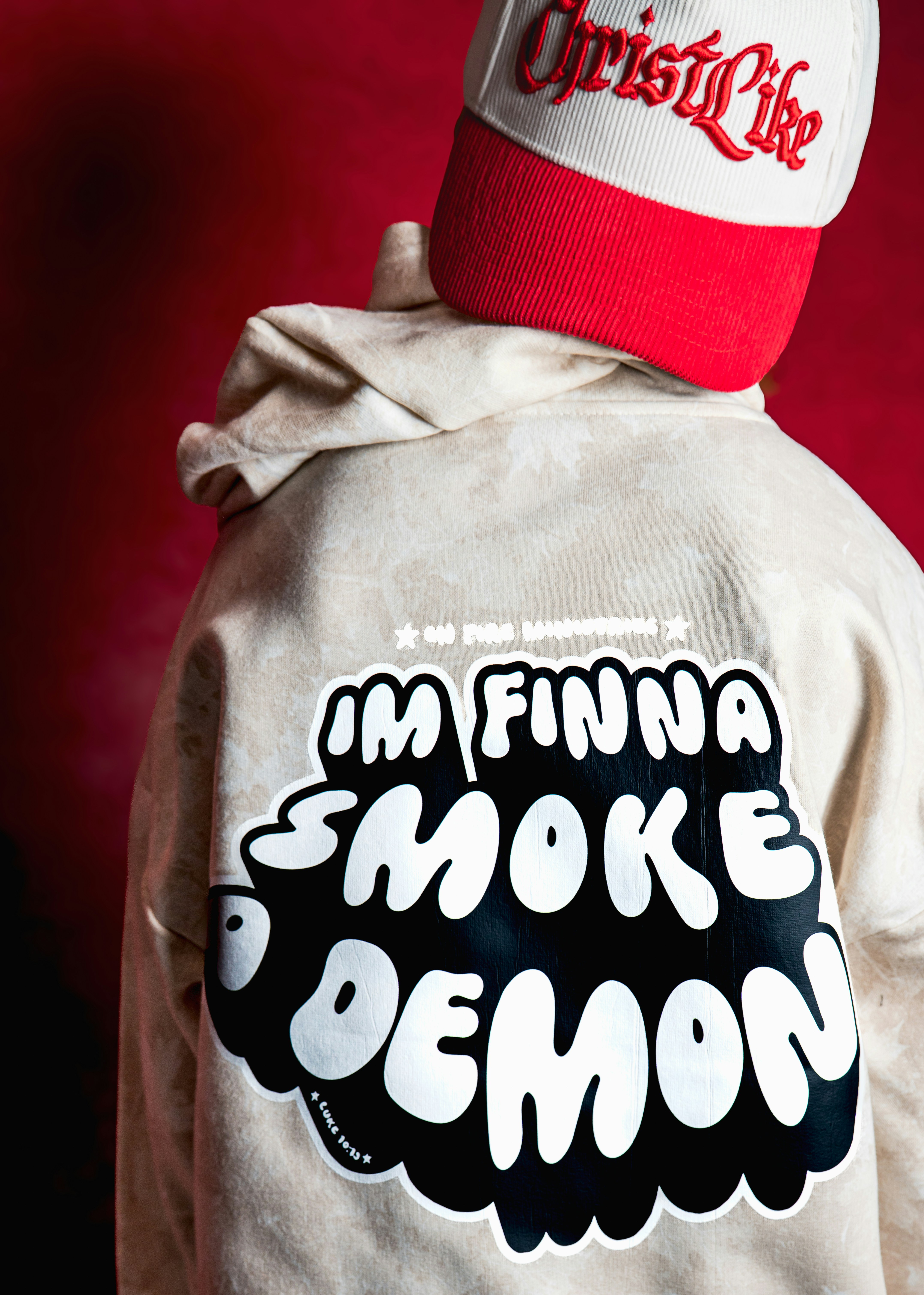 A person wearing a beige hoodie and a red and white cap against red
