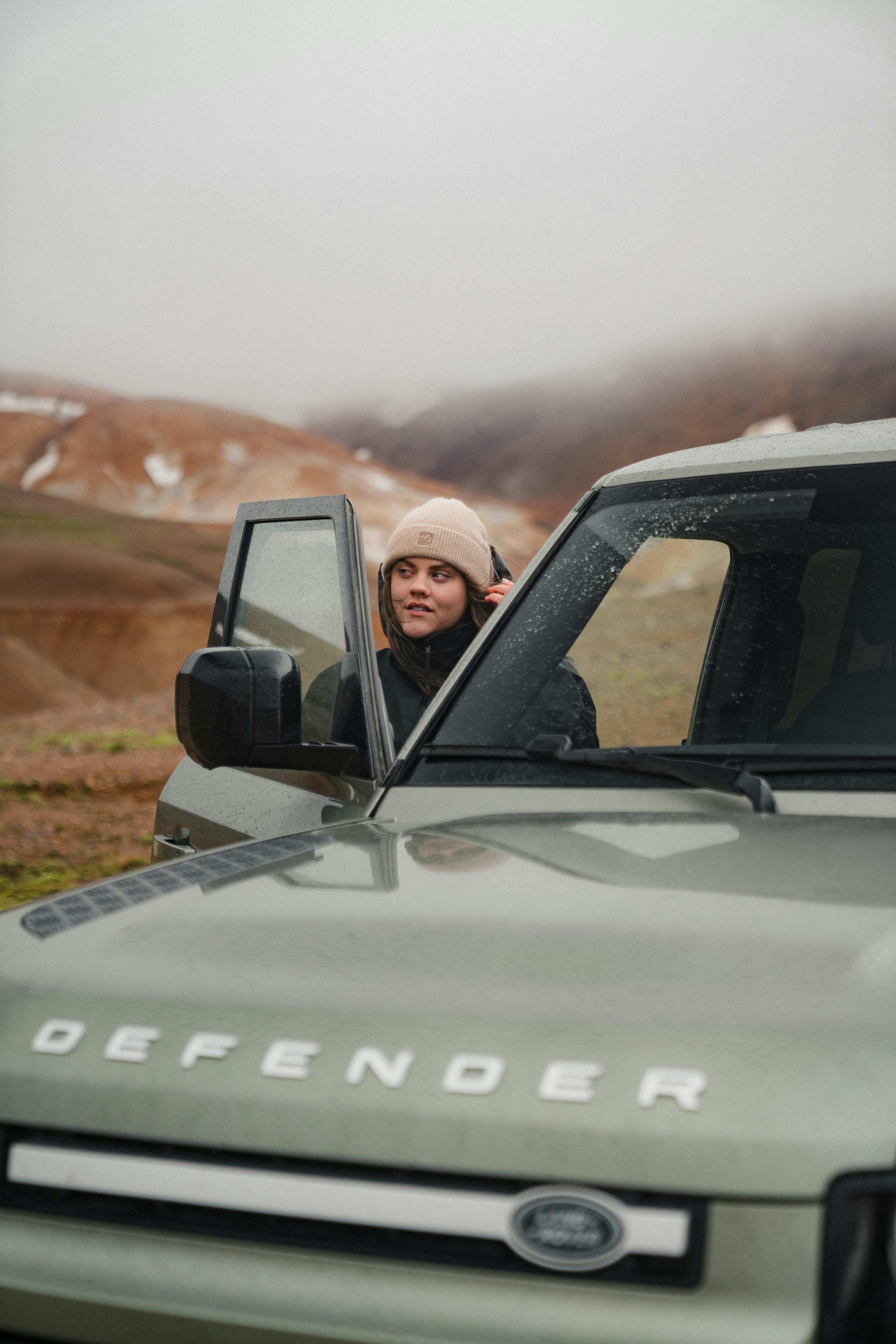 A woman in a beanie exiting a green SUV in a misty mountain landscape