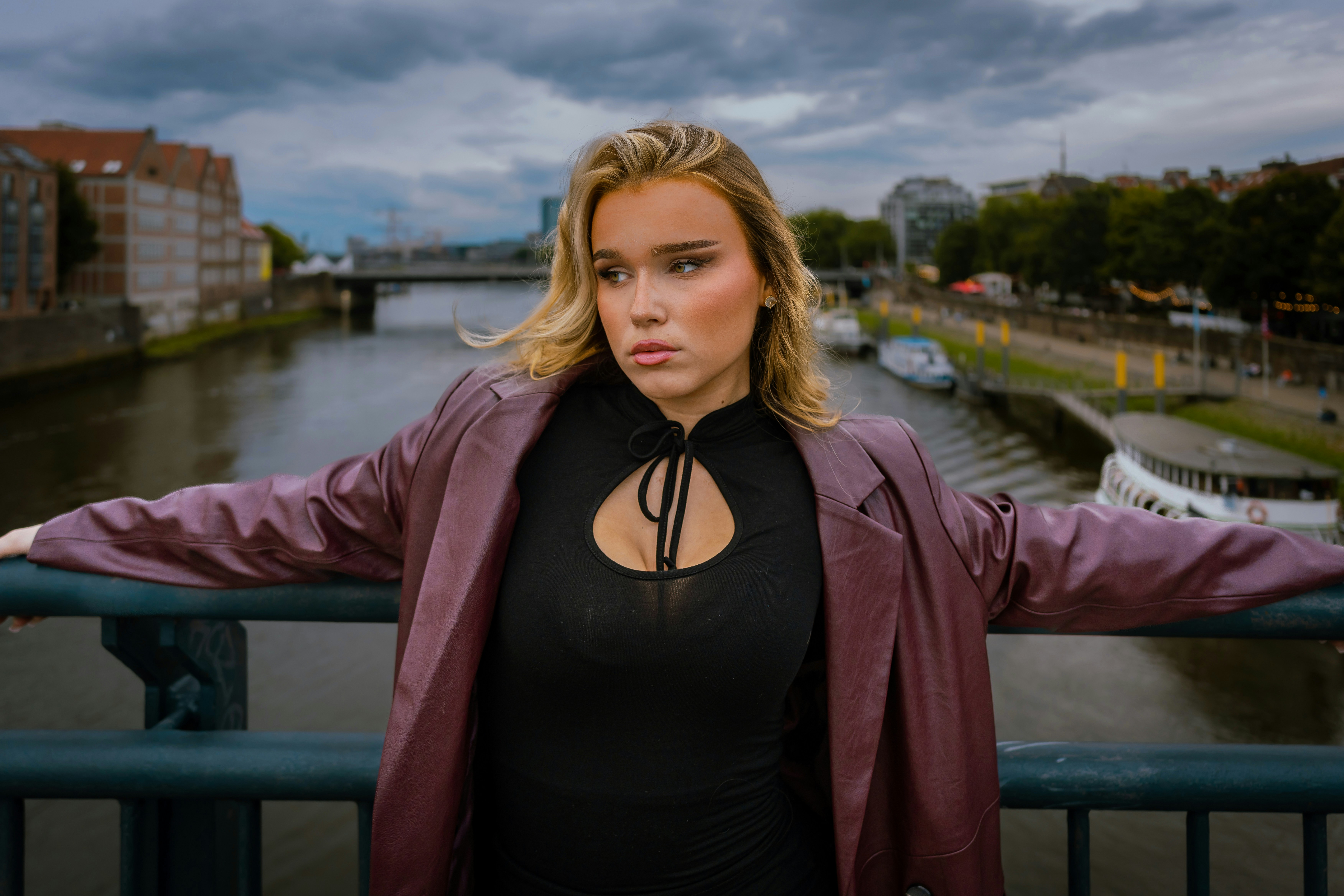 A woman in a maroon leather jacket leaning on a bridge railing over a river
