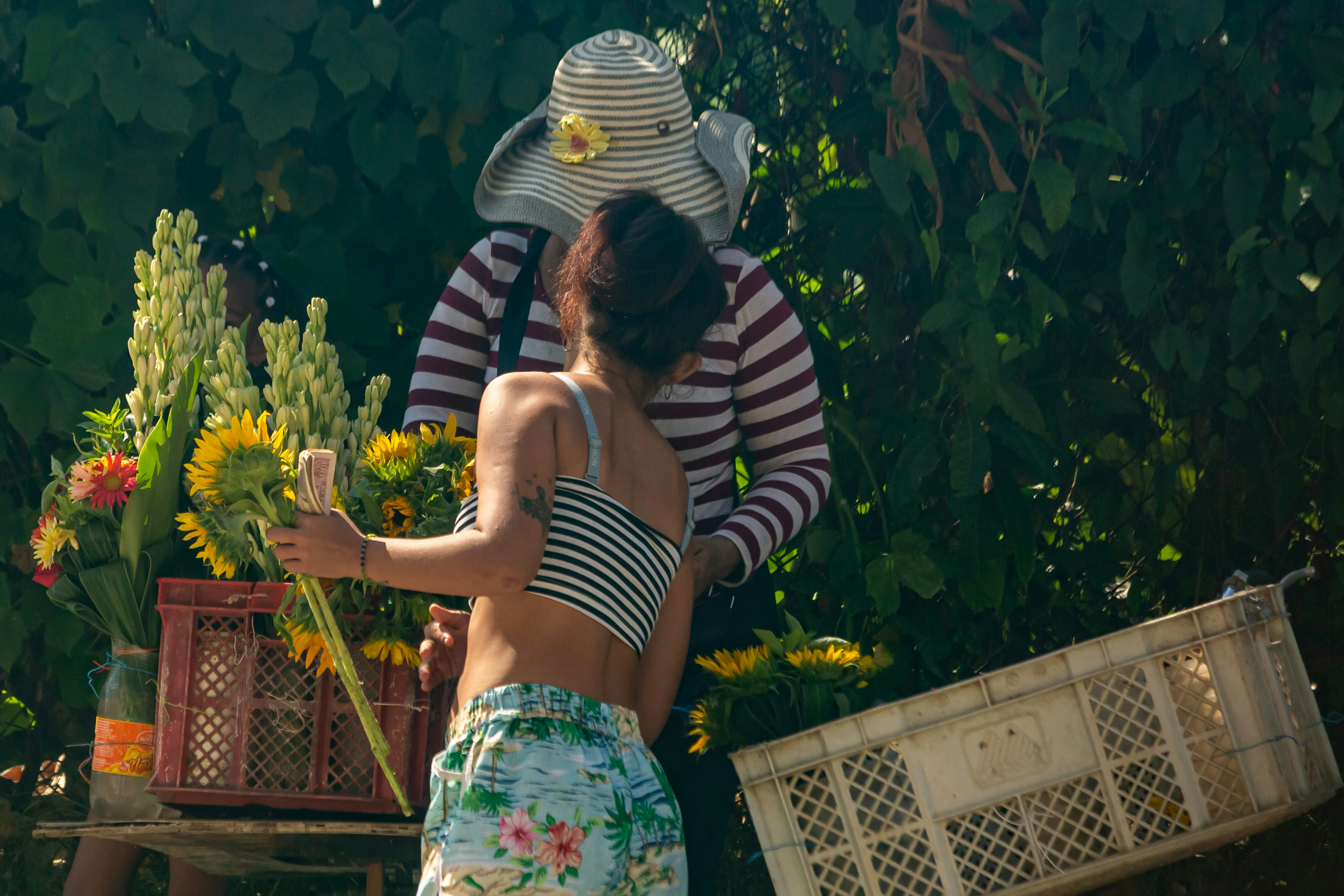A woman in a striped top and floral shorts holding sunflowers near a flower stall