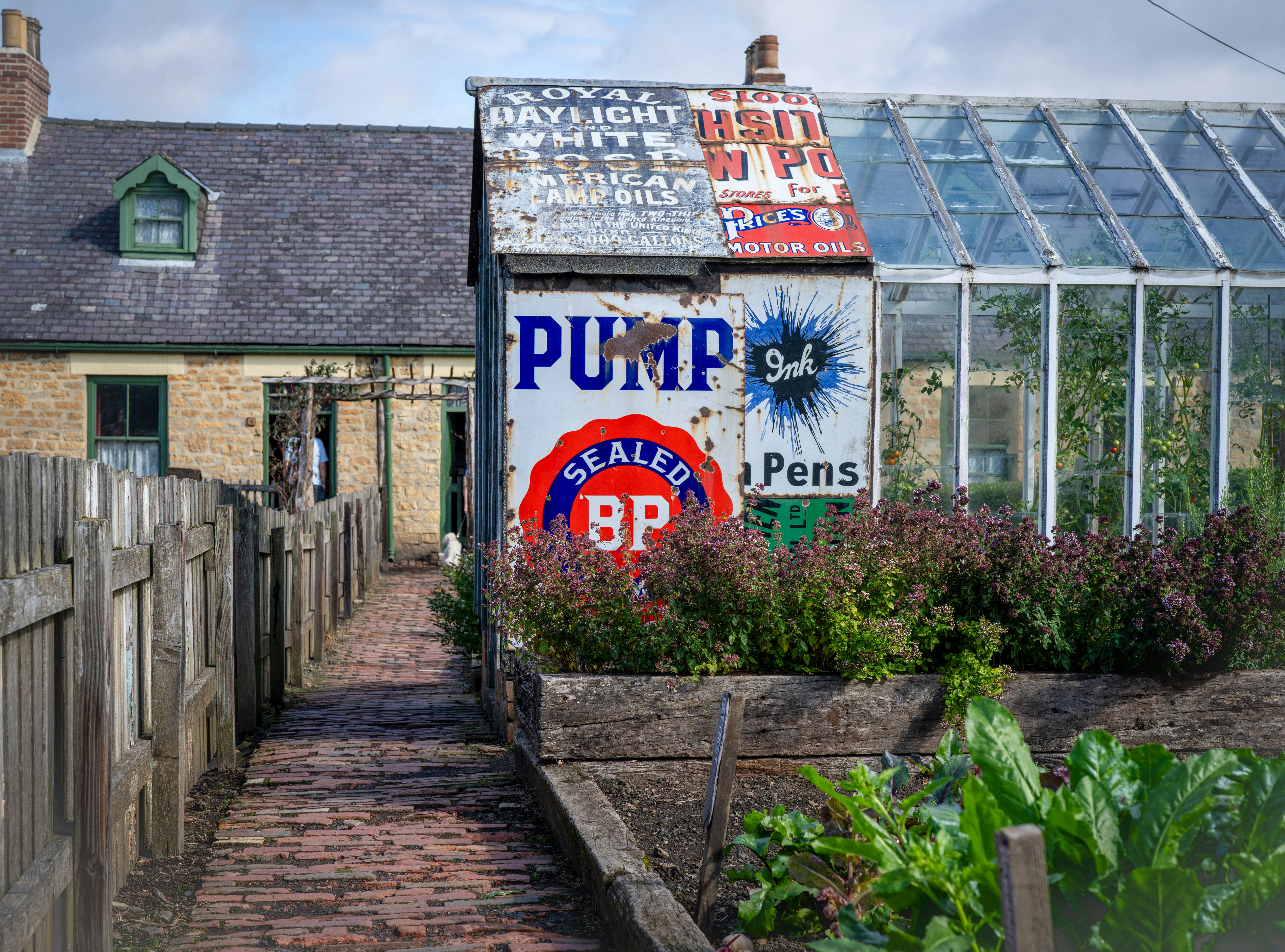 A shed covered in vintage advertising signs beside a greenhouse and brick path