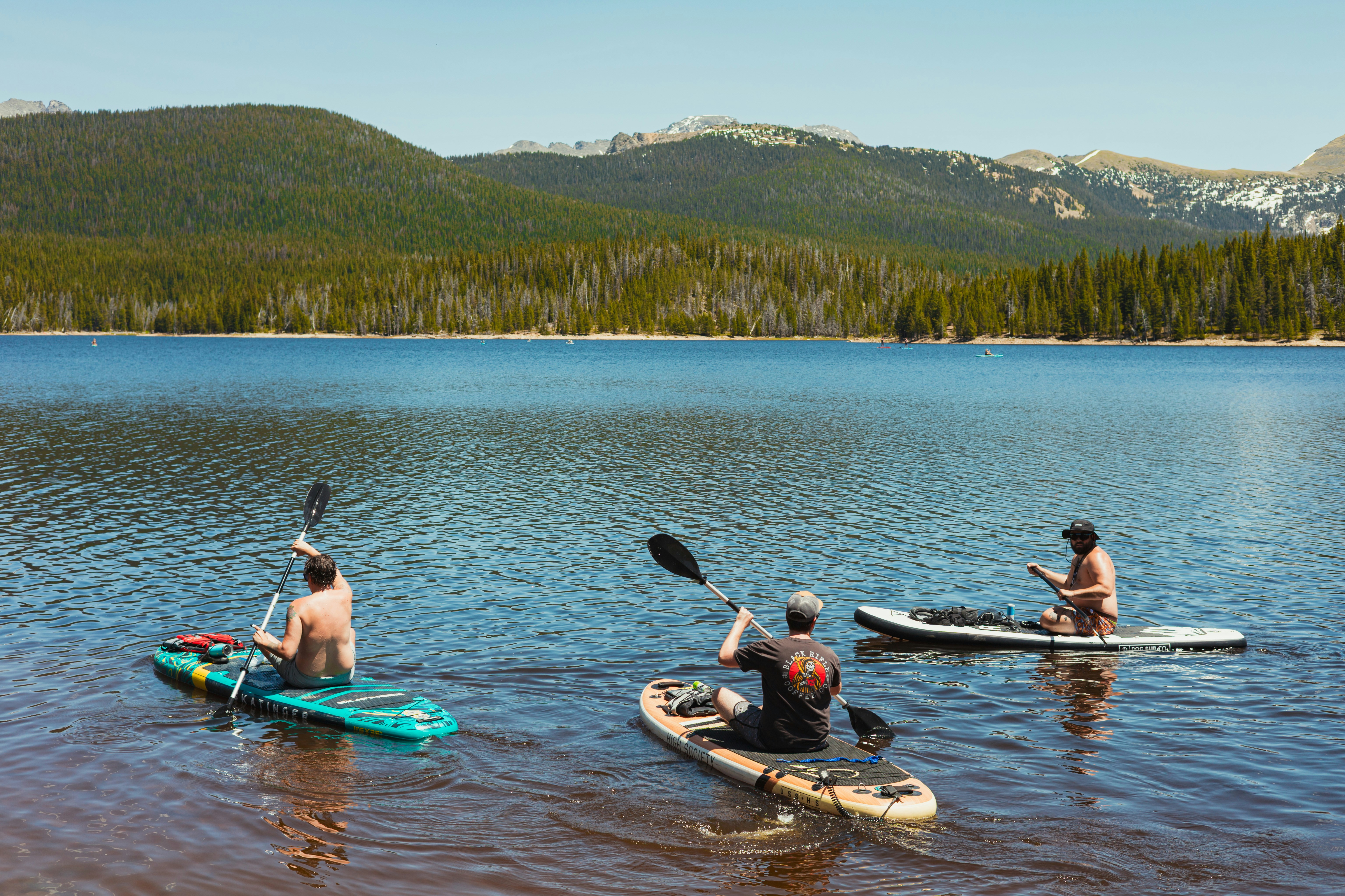 Three people paddleboarding on a blue mountain lake with pine trees and peaks