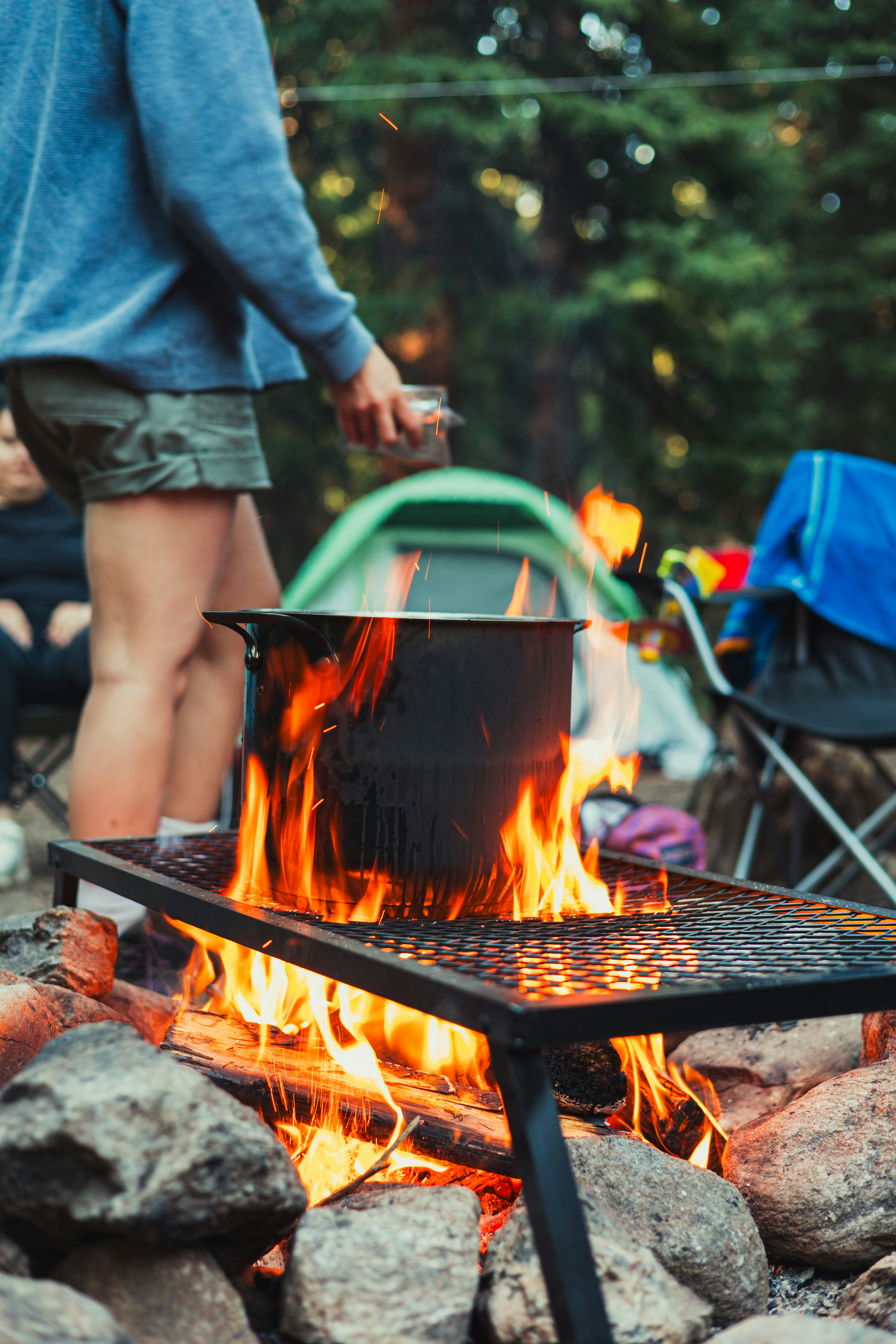 Pot of food heating on a metal grate over a campfire ringed with stones, a camper standing beside it and tents and folding chairs behind