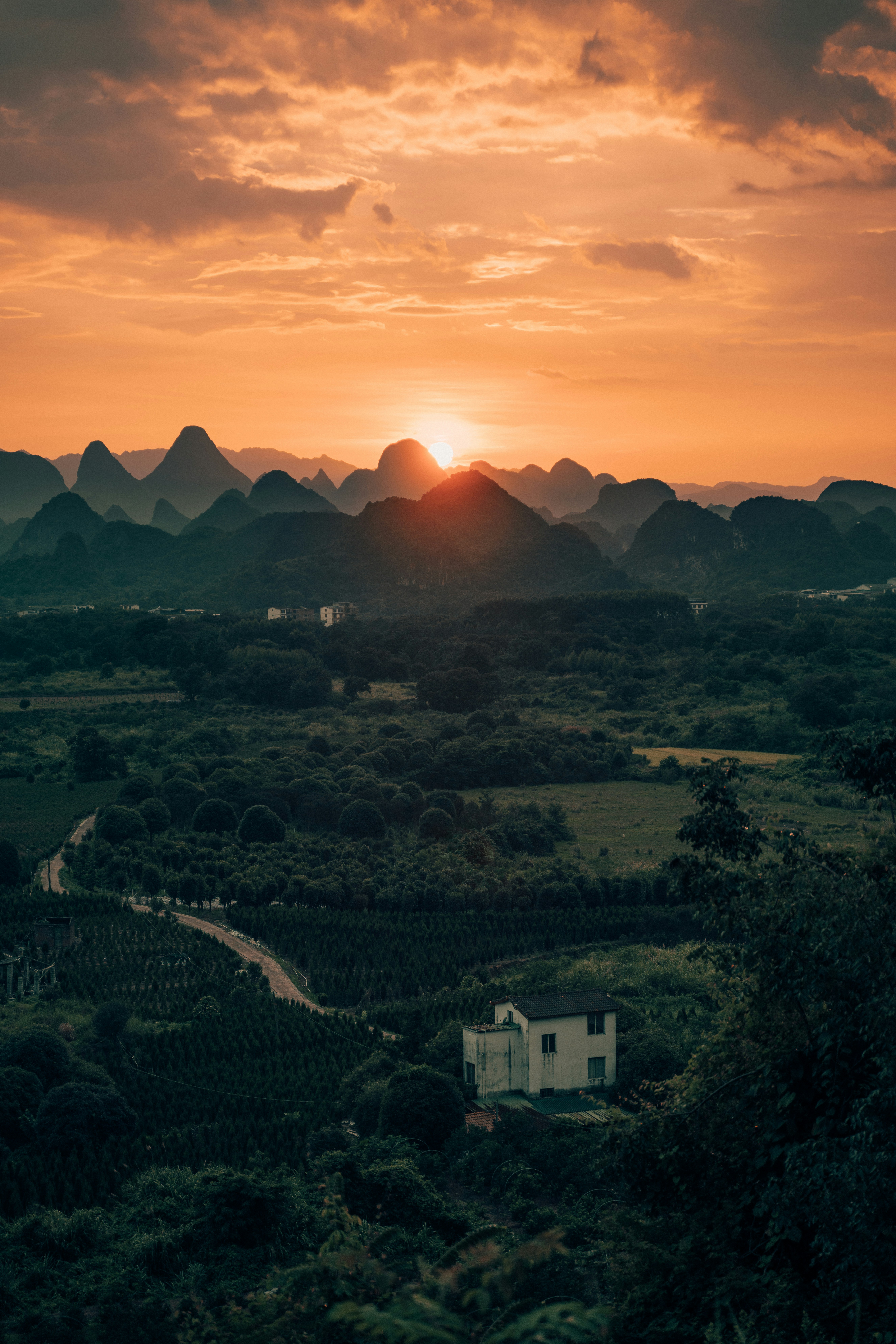 A small house in a valley below karst mountains under a warm orange sky