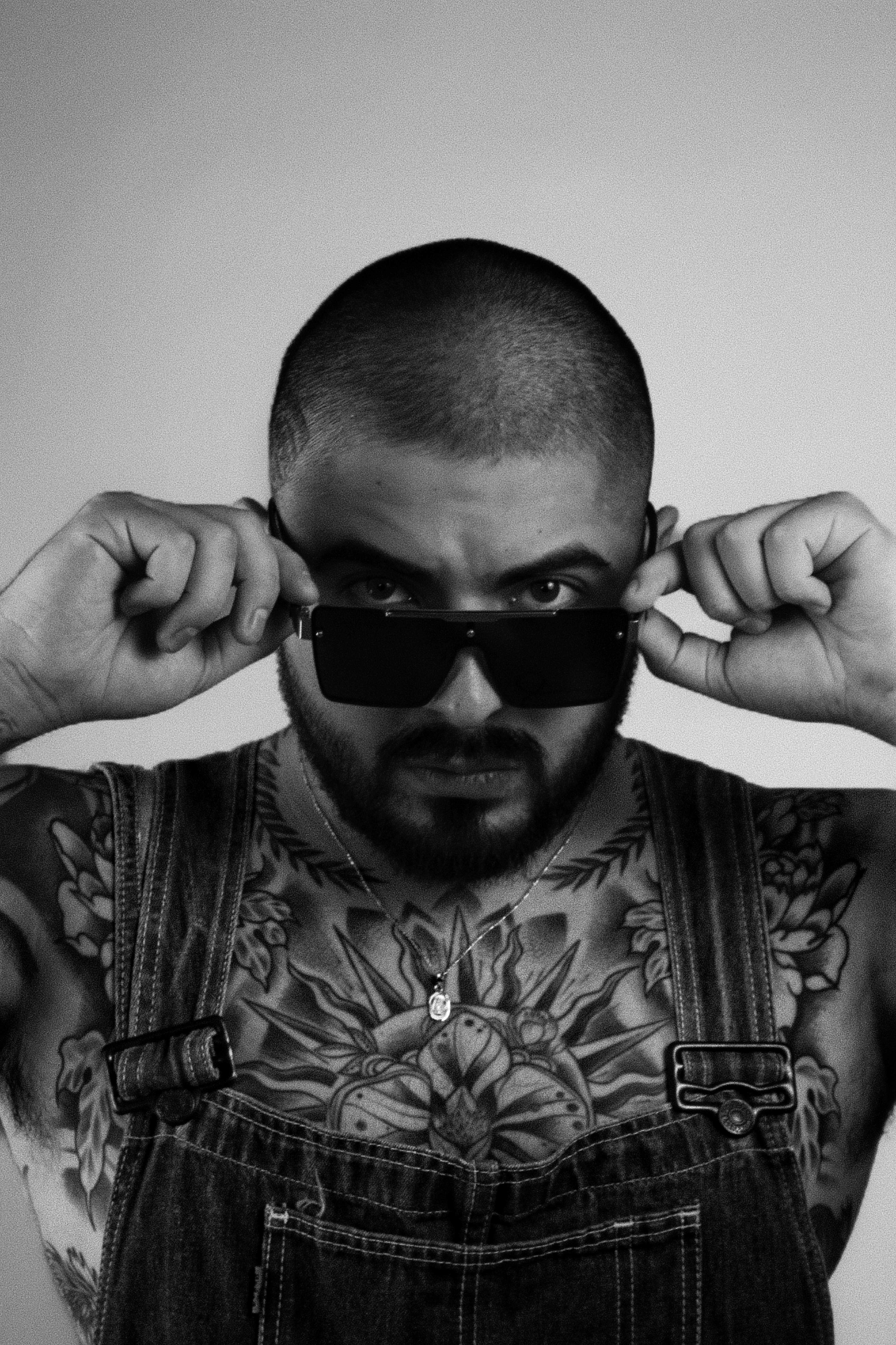 A man with a shaved head and tattoos wearing sunglasses and denim overalls