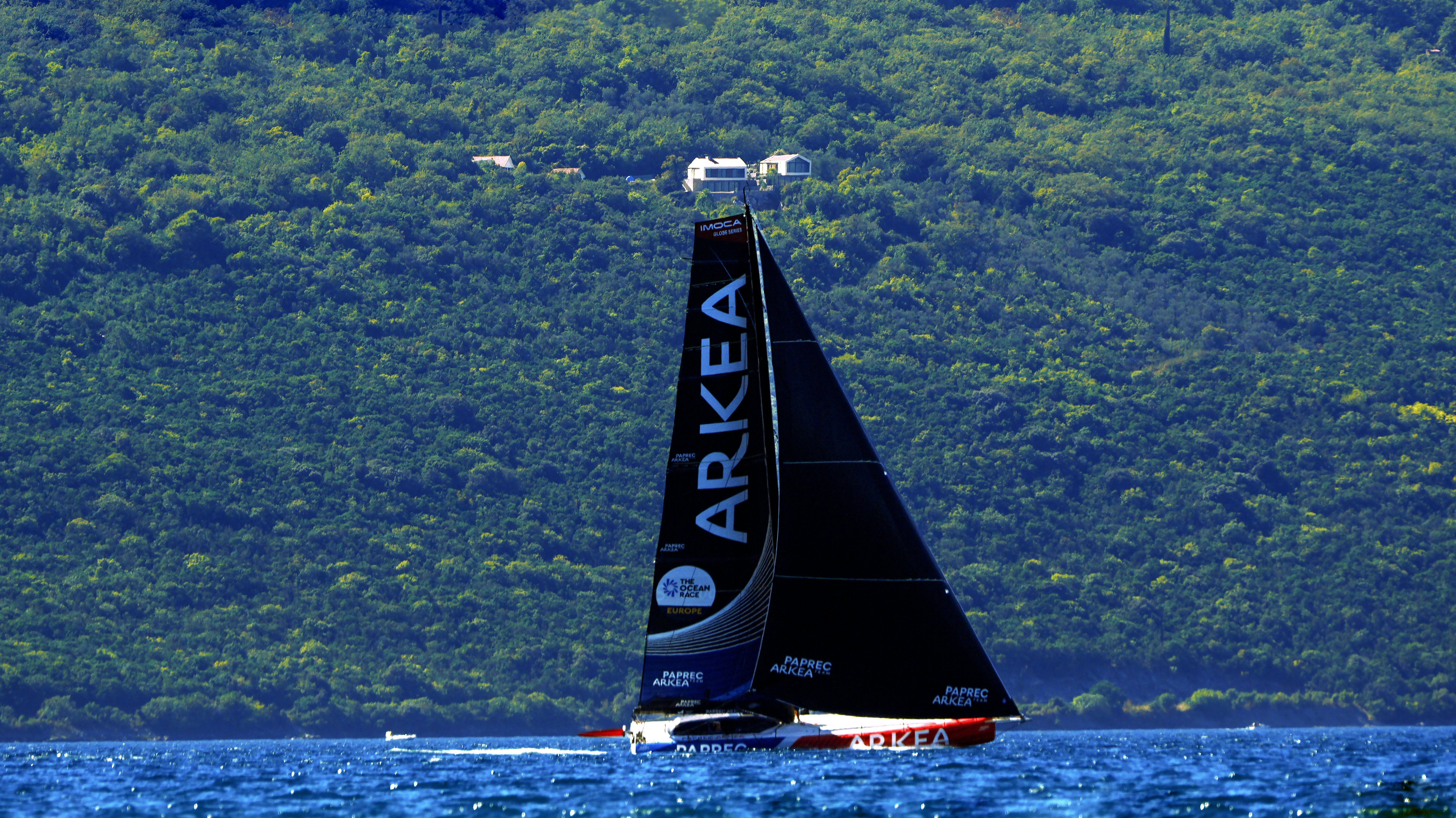 A racing sailboat with a black sail on blue water before a forested mountain