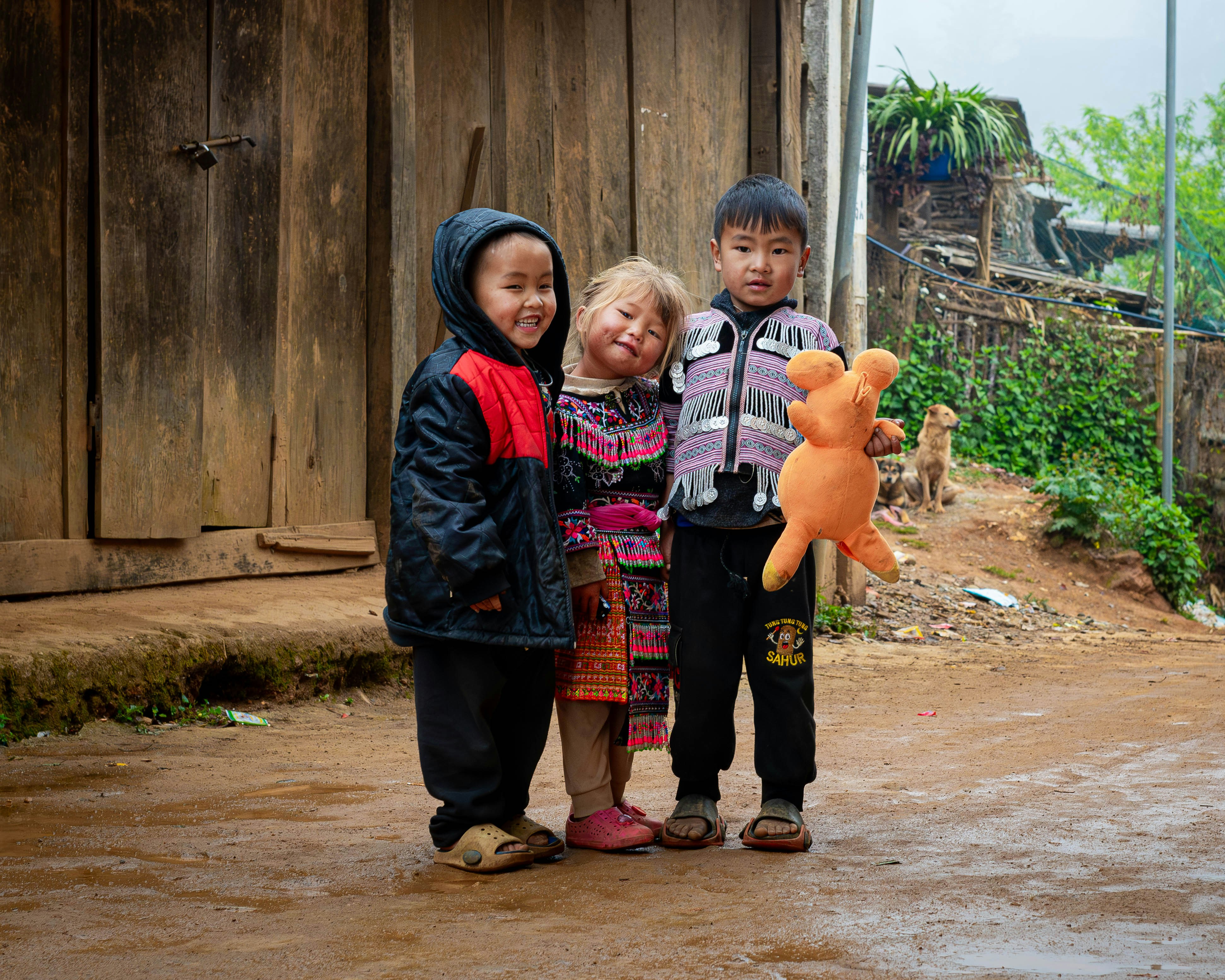 Three children standing on a dirt path in front of a wooden building