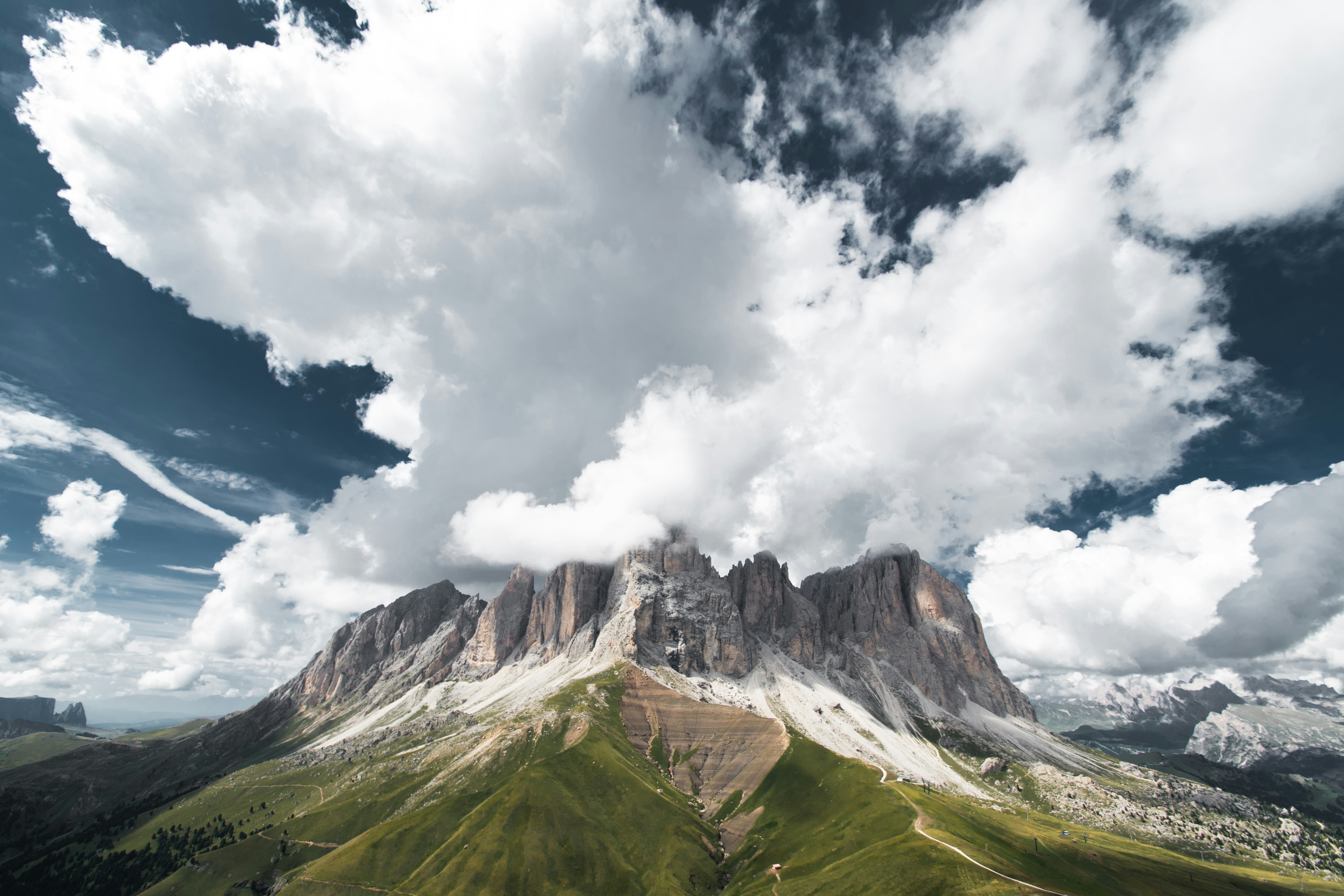 Gezackte Berggipfel, die unter einer massiven weißen Wolkenformation über grüne Hänge ragen