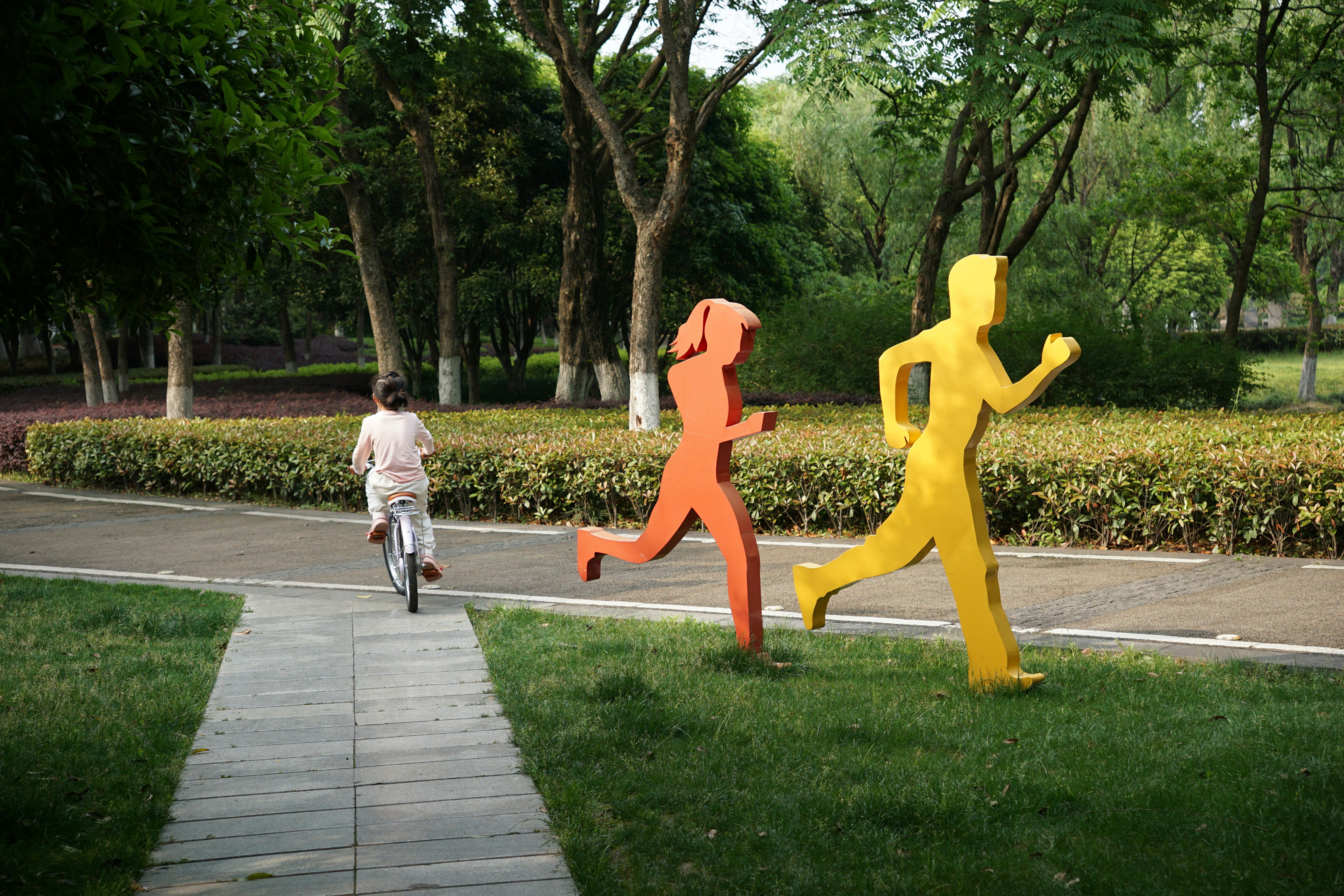 A child riding a bicycle past orange and yellow runner sculptures in a park