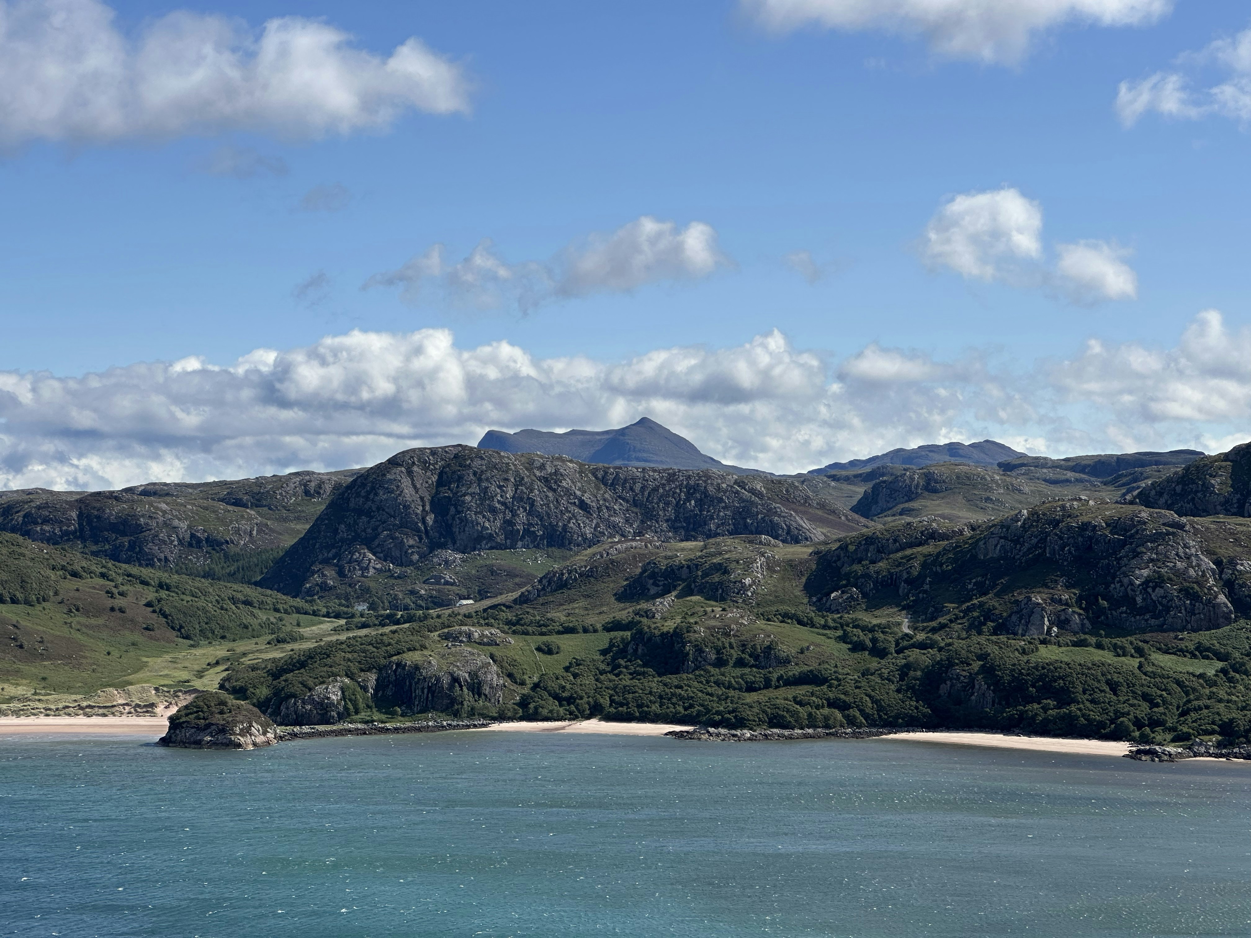A sandy beach below rocky hills and green slopes under a blue sky