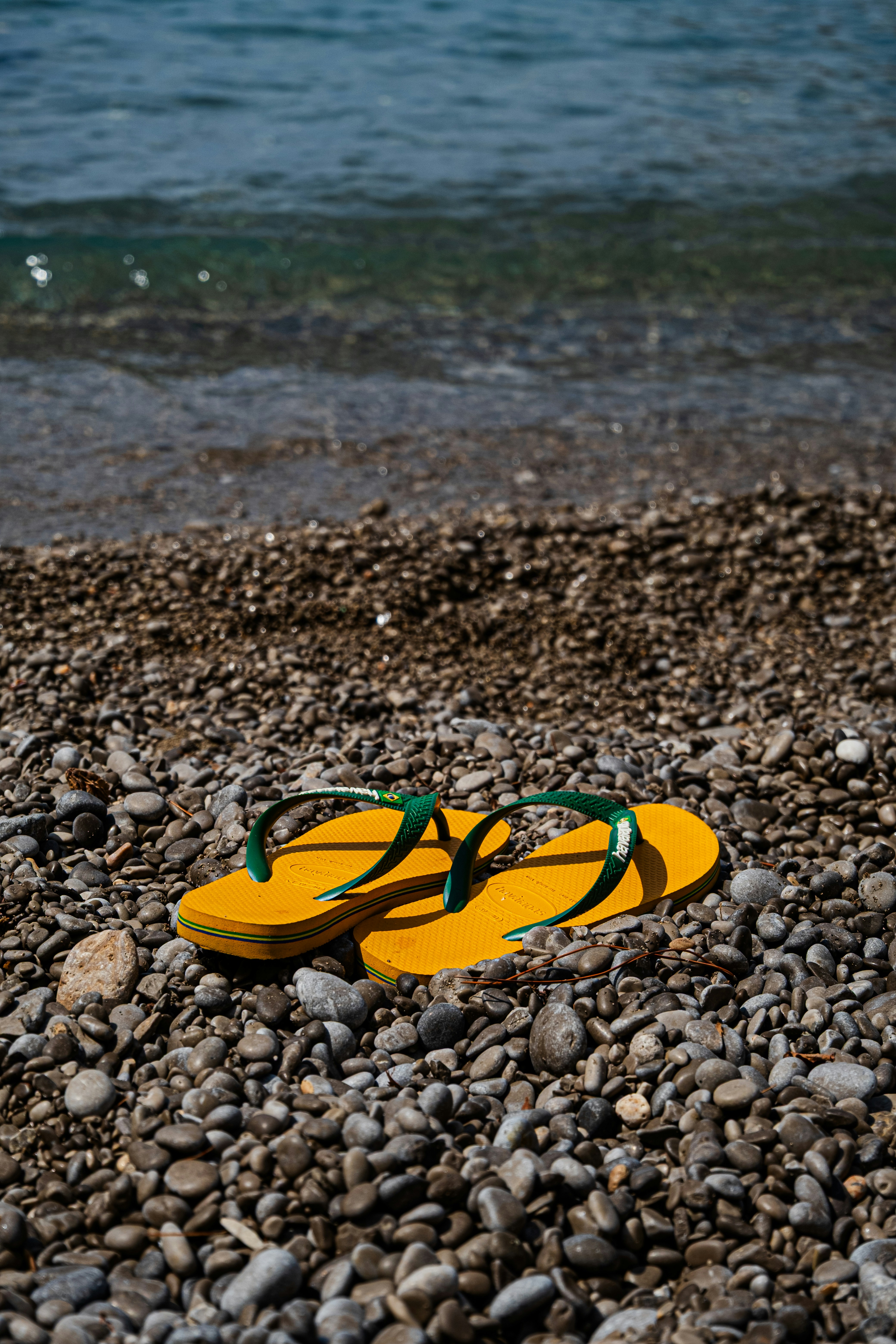 Yellow flip-flops with green straps on a pebble beach near the water