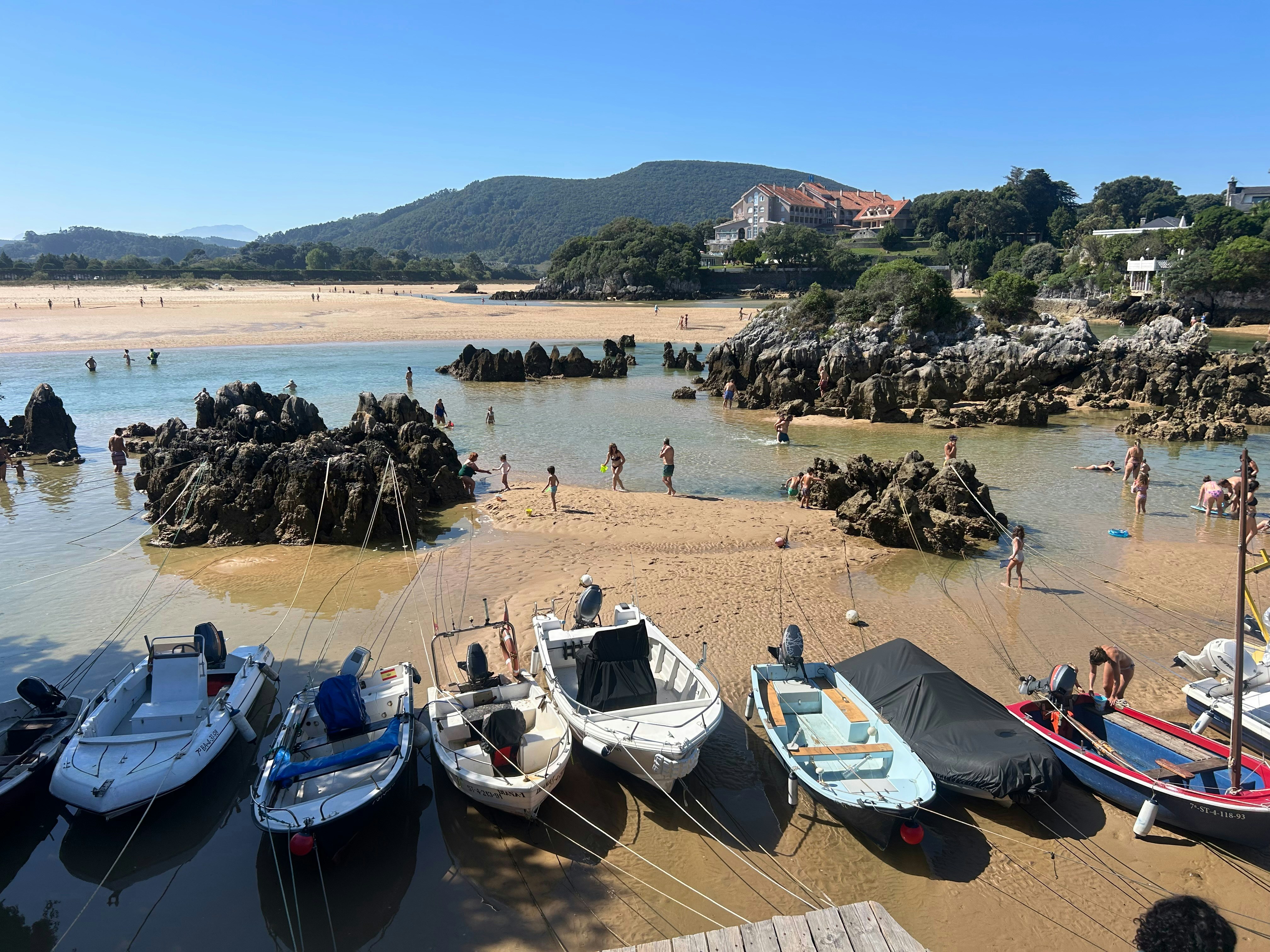 Small motorboats moored on a sandy beach with people swimming among rocky outcrops