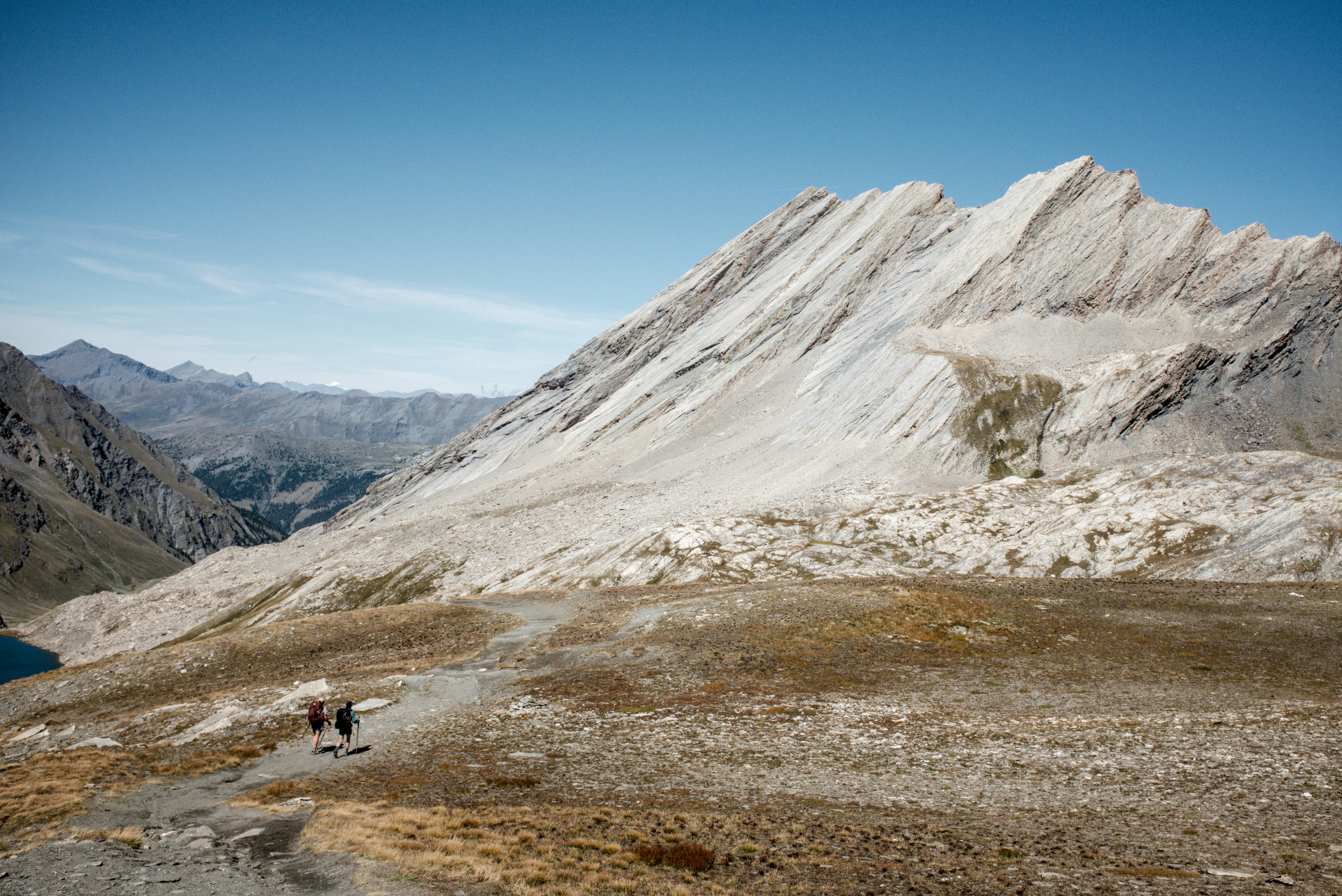 Two hikers with backpacks walking on a trail below a large rocky mountain